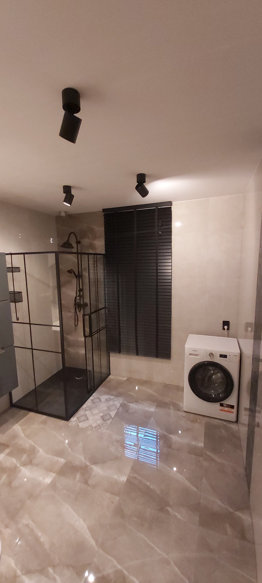 Wykończona łazienka z kabiną prysznicową typu walk-in z czarnymi profilami, marmurową podłogą i białą pralką pod oknem z żaluzjami.
