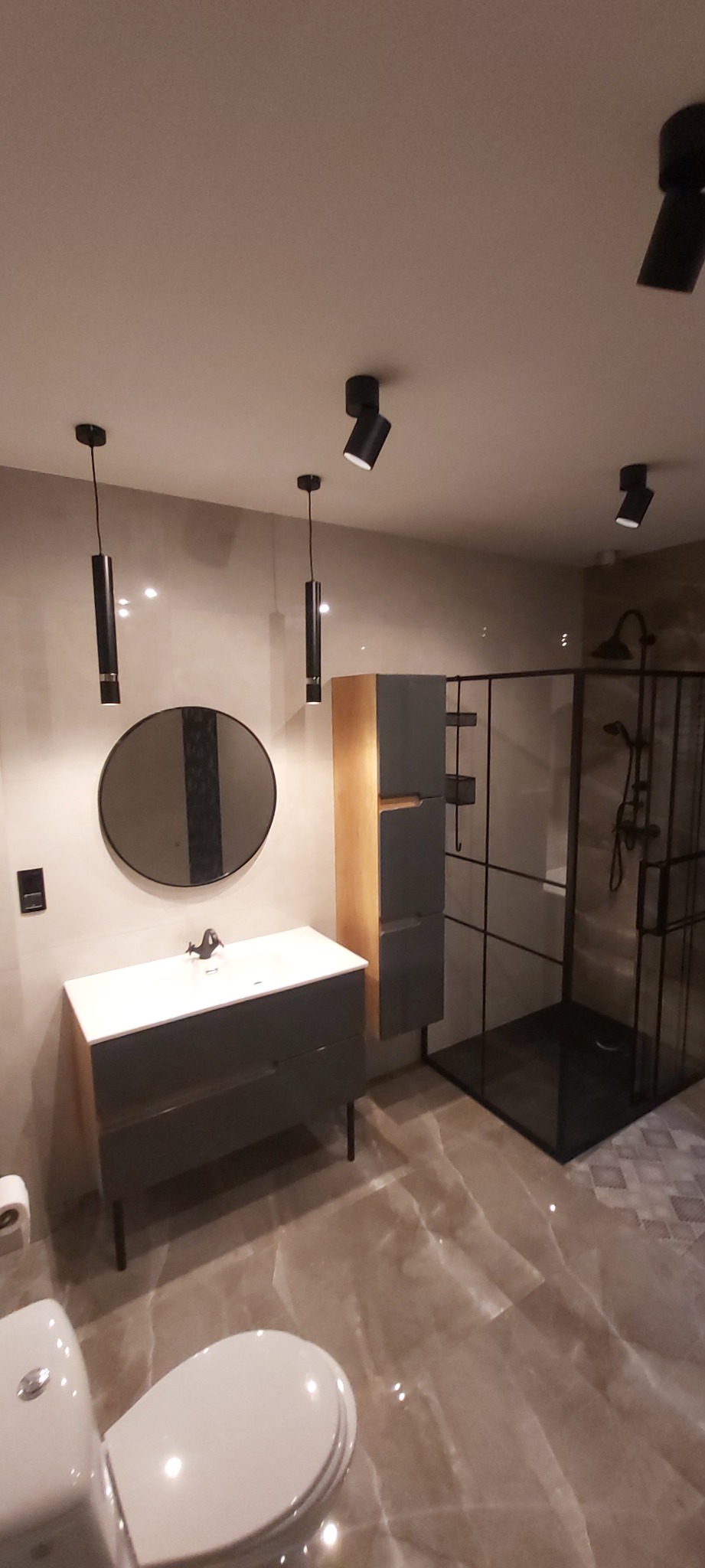 Nowoczesna łazienka z umywalką w szarej szafce, okrągłym lustrem z czarnym akcentem, kabiną prysznicową typu walk-in z czarnymi ramami i deszczownicą, oraz punktowym oświetleniem sufitowym.