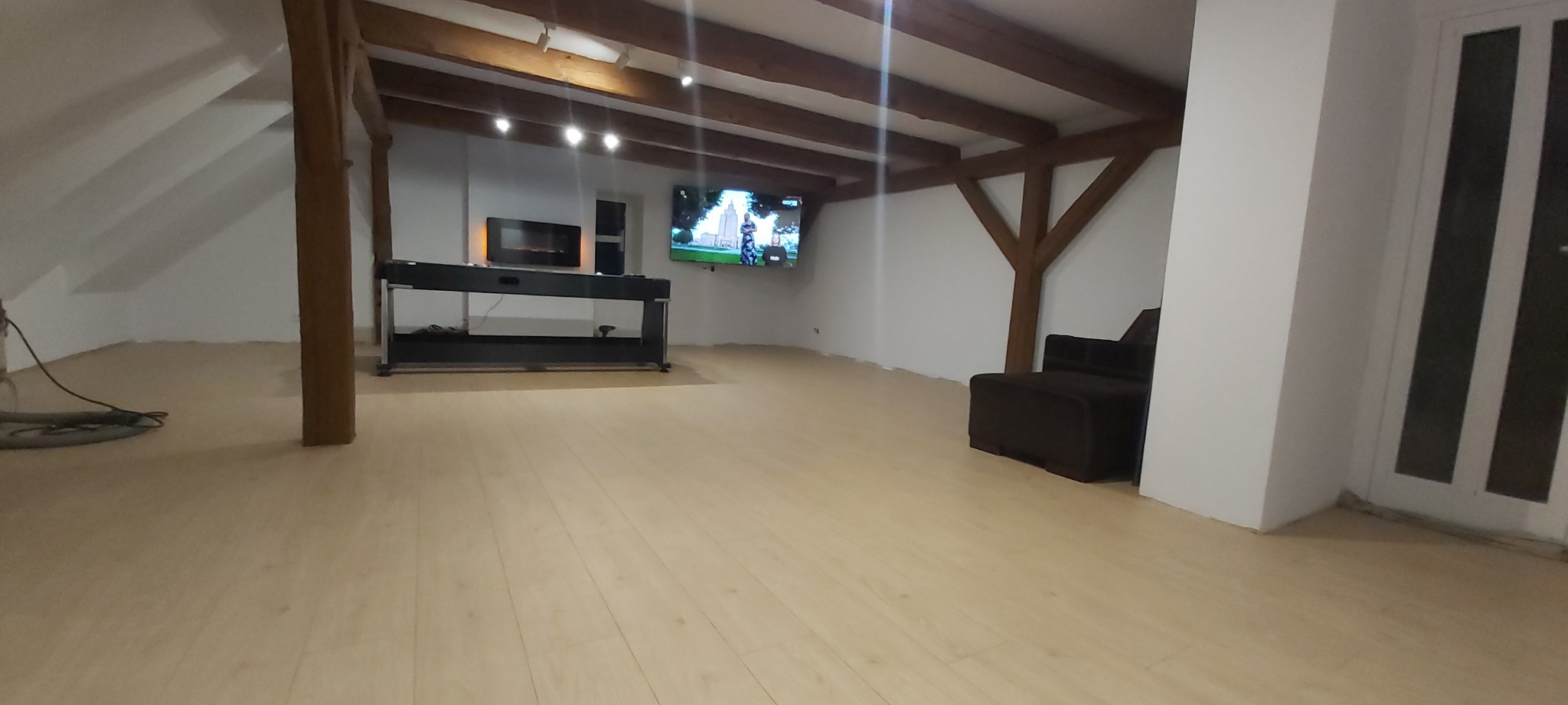 Jasne panele podłogowe w pokoju na poddaszu z drewnianymi belkami stropowymi i telewizorem na ścianie, widoczny odkurzacz przemysłowy.