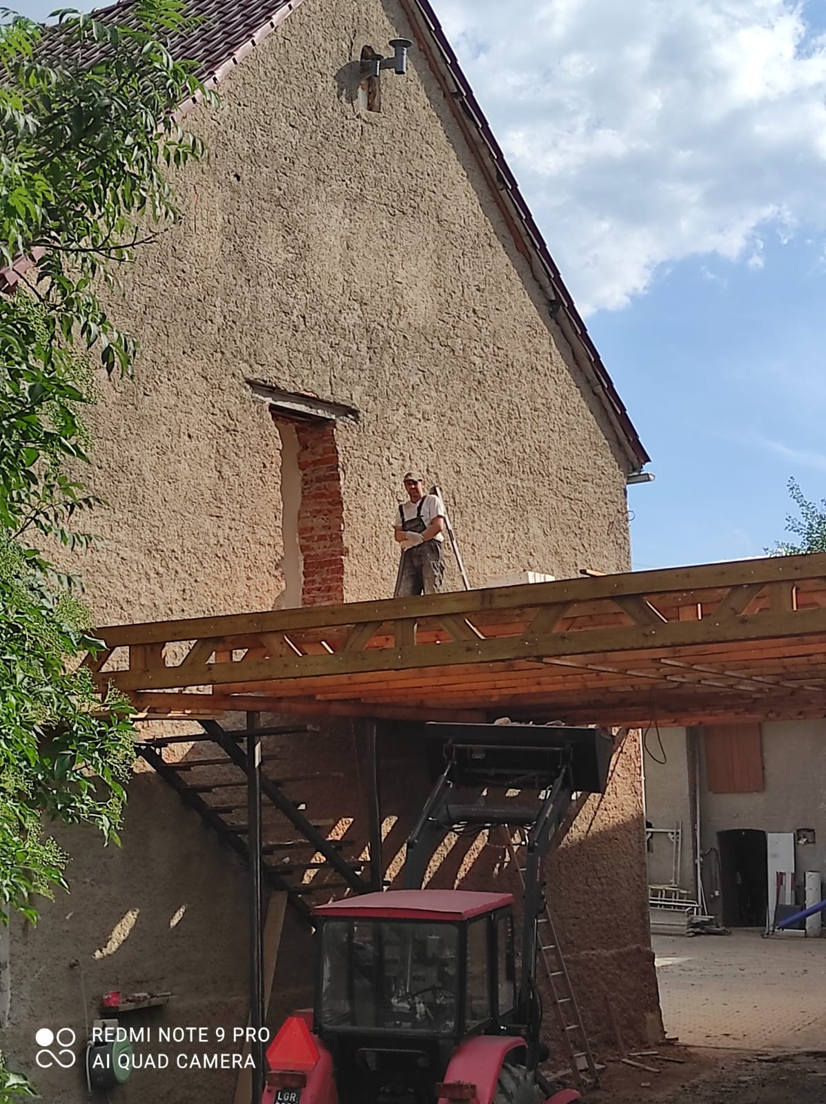 Mężczyzna w kombinezonie i okularach, stojący na drewnianej platformie przy budynku z surową elewacją, w tle czerwony traktor z podnośnikiem, prawdopodobnie podczas prac remontowych.