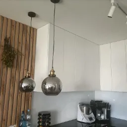 Dwa szklane klosze lamp wiszących w kuchni z białymi szafkami i drewnianą ścianą, widoczne także reflektory sufitowe, ekspres do kawy i Thermomix.