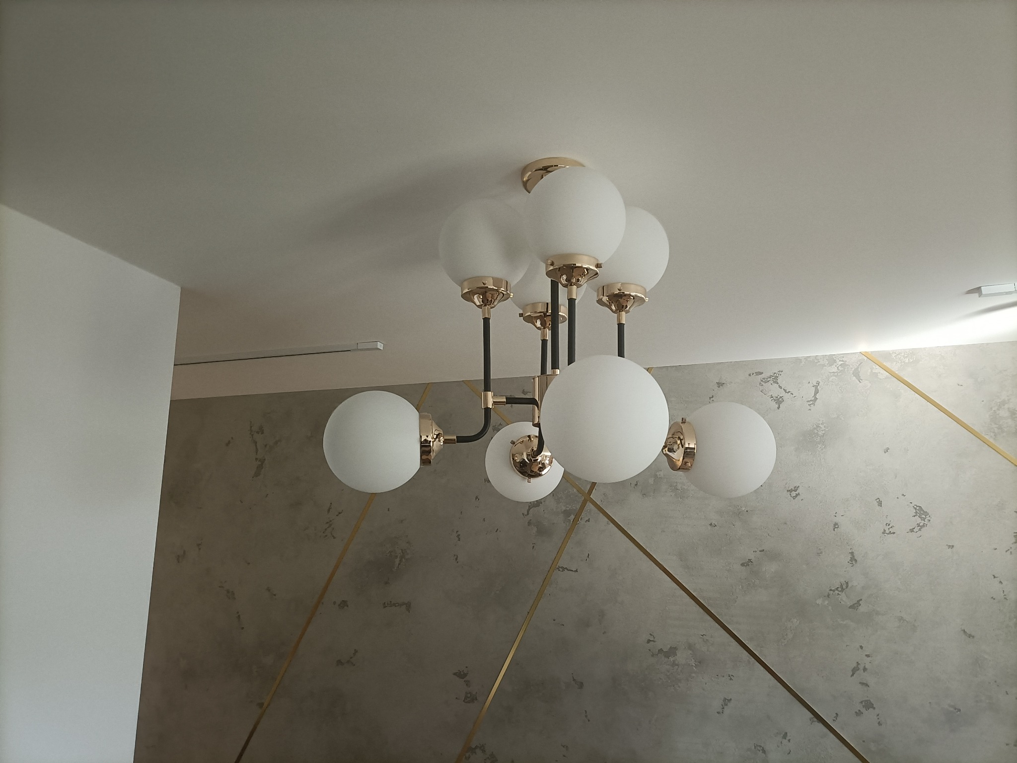 Nowoczesny żyrandol z białymi kulami i złotymi detalami zamontowany na suficie w Minkowicach, widoczna dekoracyjna ściana z geometrycznym wzorem.