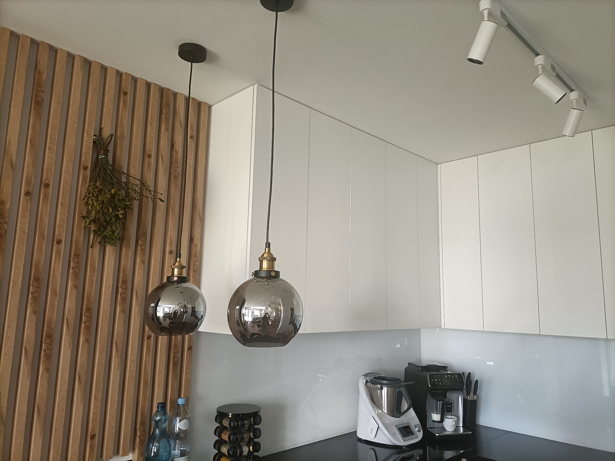 Dwa szklane klosze lamp wiszących w kuchni z białymi szafkami i drewnianą ścianą, widoczne także reflektory sufitowe, ekspres do kawy i Thermomix.