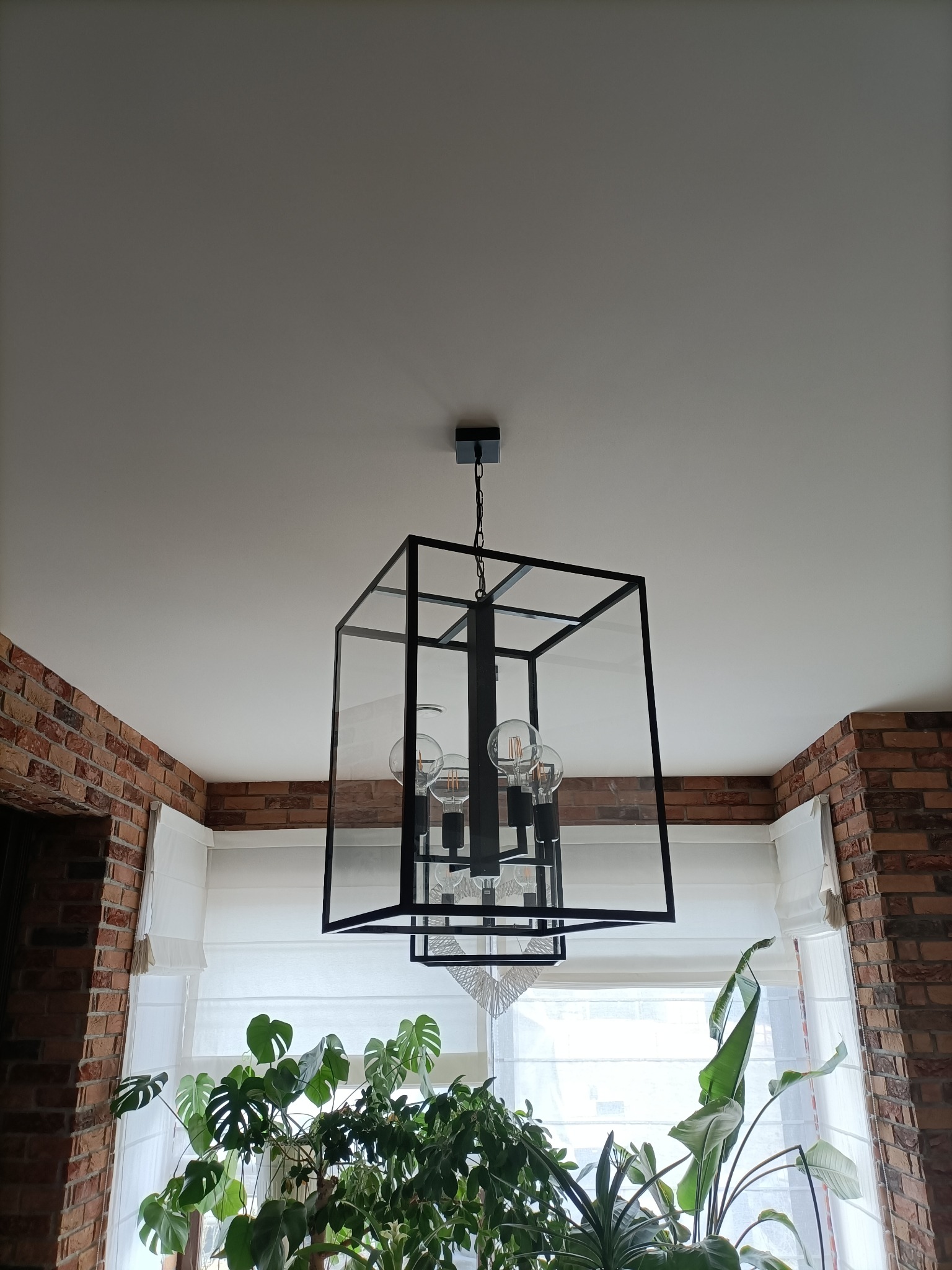 Czarna, geometryczna lampa wisząca w stylu industrialnym, z żarówkami Edisona, na tle ceglanej ściany, białych rolet rzymskich i roślin doniczkowych.
