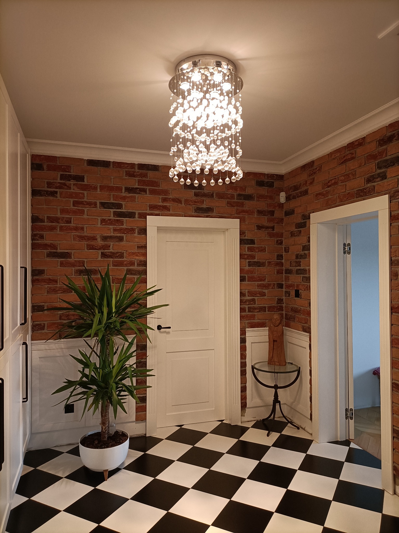 Kryształowy żyrandol sufitowy w przestronnym holu z ceglaną ścianą, białą stolarką drzwiową i białą szafą w zabudowie, na podłodze czarno-biała szachownica.