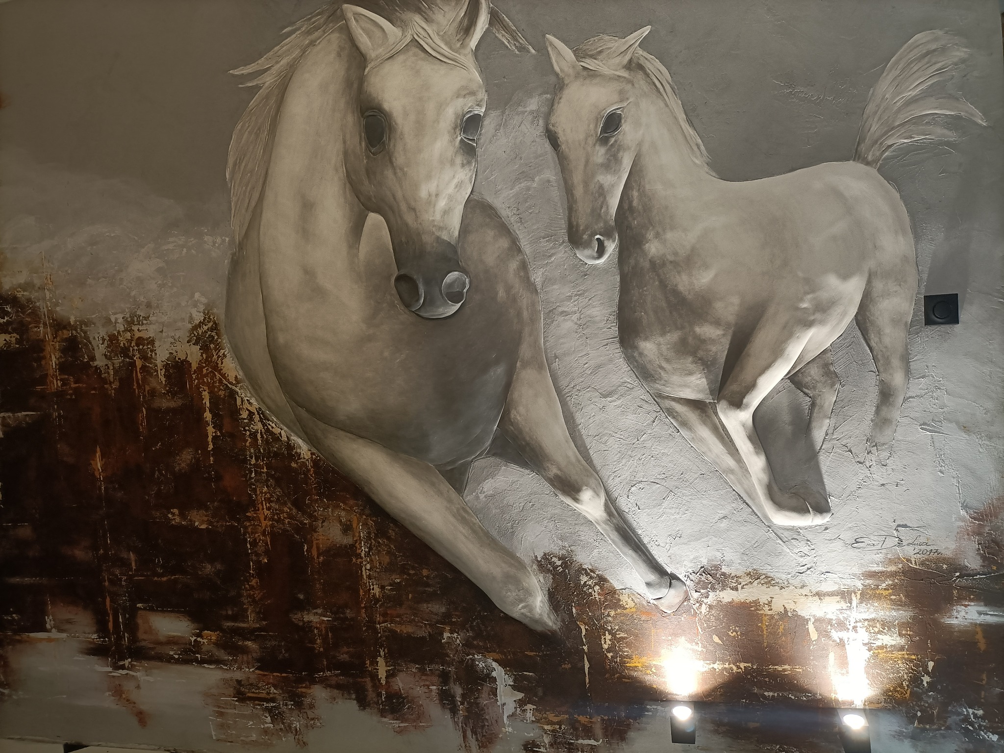 Szara ściana z fakturą i ręcznie malowanym obrazem dwóch galopujących koni w odcieniach szarości i brązu, oświetlona od dołu halogenami.