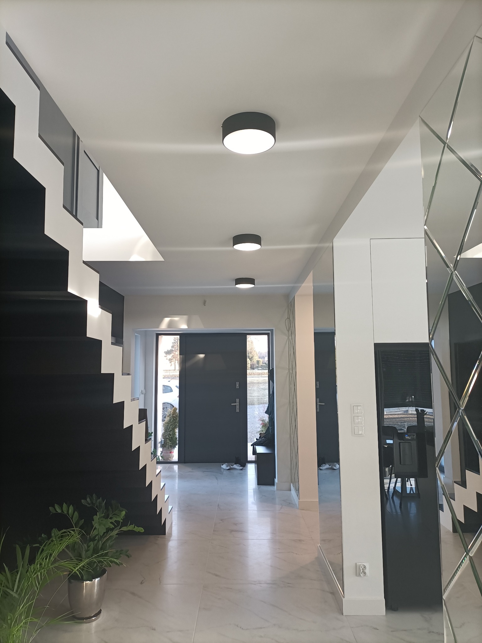 Nowoczesny przedpokój z czarno-białymi schodami, marmurową podłogą i okrągłymi lampami sufitowymi, odbijający się w lustrzanej ścianie.