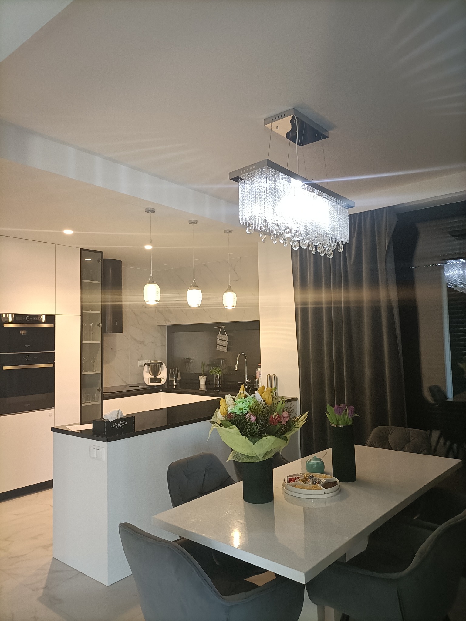 Nowoczesna kuchnia z wyspą i białymi szafkami, oświetlona kryształowym żyrandolem i trzema wiszącymi lampami, marmurowa ściana w tle, stół z kwiatami na pierwszym planie.