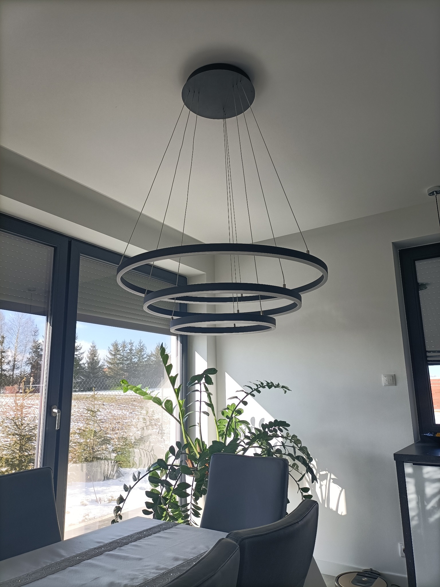 Nowoczesny, trzypoziomowy, okrągły żyrandol LED w szaro-czarnej kolorystyce, zawieszony nad stołem w jasnym wnętrzu z widokiem na zaśnieżony ogród przez duże okno.