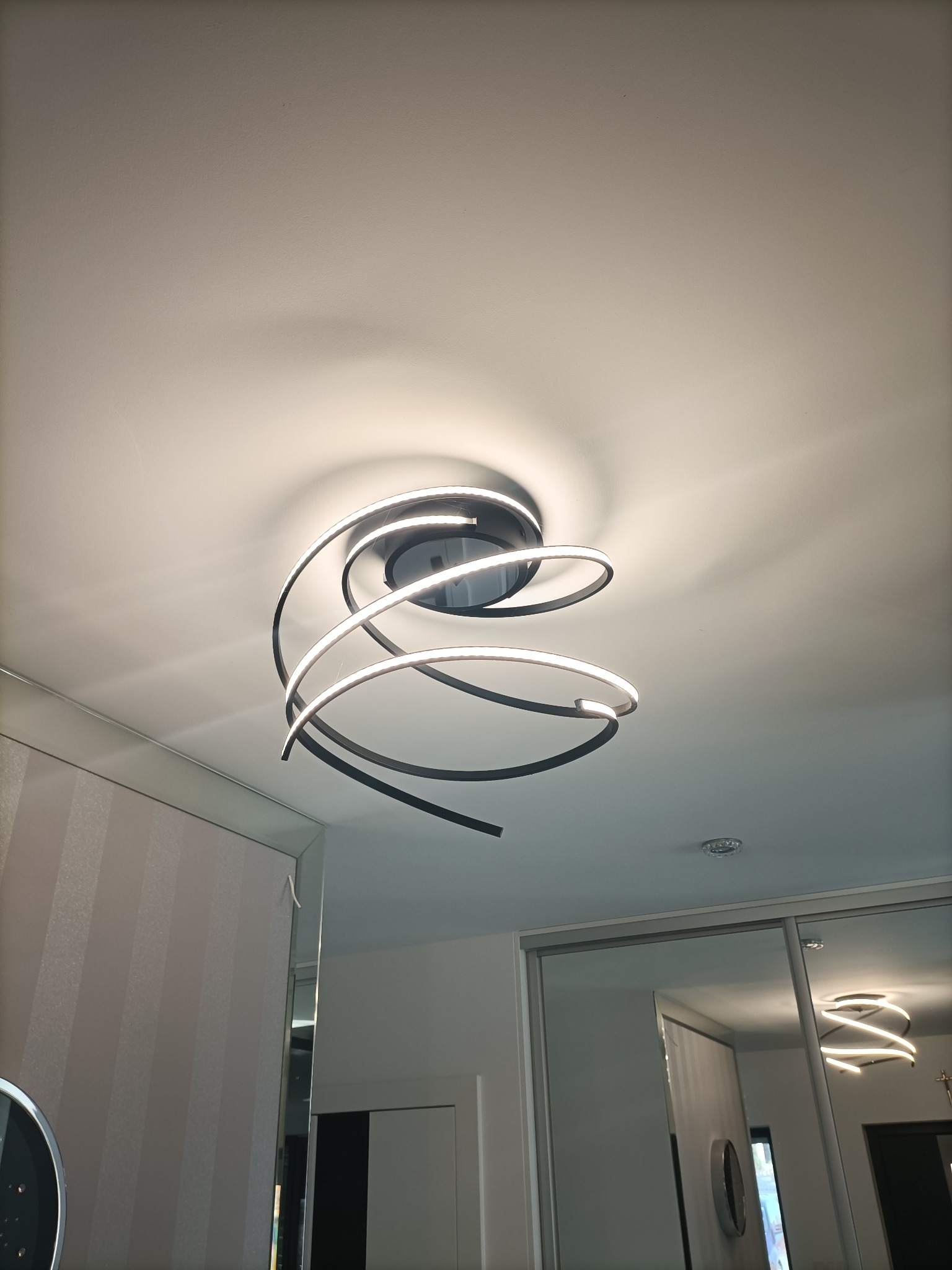 Nowoczesna lampa sufitowa LED w kształcie spirali, z czarną obudową i białym światłem, zamontowana na białym suficie w pokoju z przesuwanymi drzwiami szafy z lustrami.