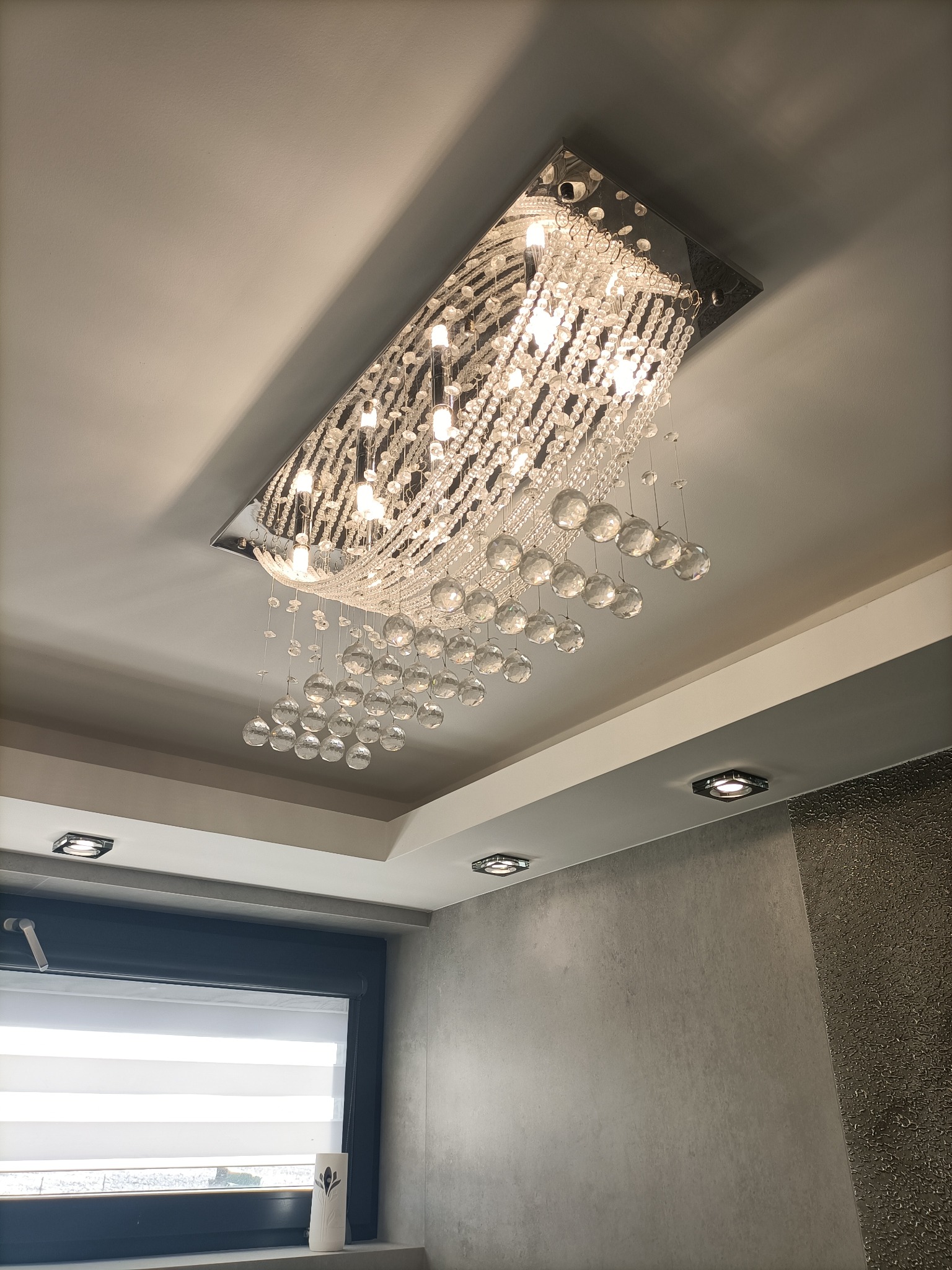 Kryształowy żyrandol w kształcie prostokąta, zamontowany na suficie, oświetlający wnętrze z szarą ścianą i oknem z roletami.