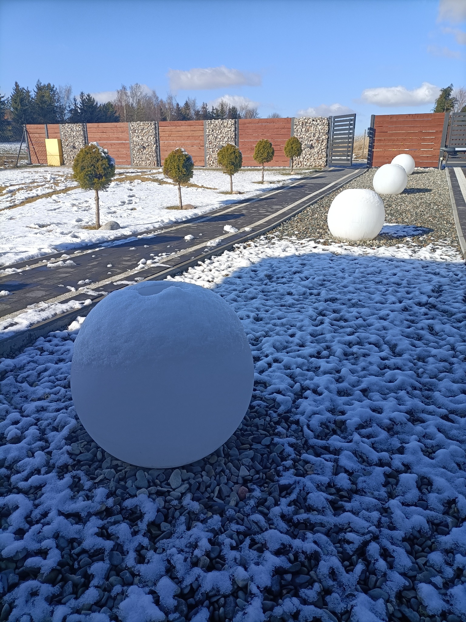 Ogród zimą z kulistymi lampami ogrodowymi pokrytymi śniegiem, kamienny chodnik i ozdobne ogrodzenie z gabionami w tle.