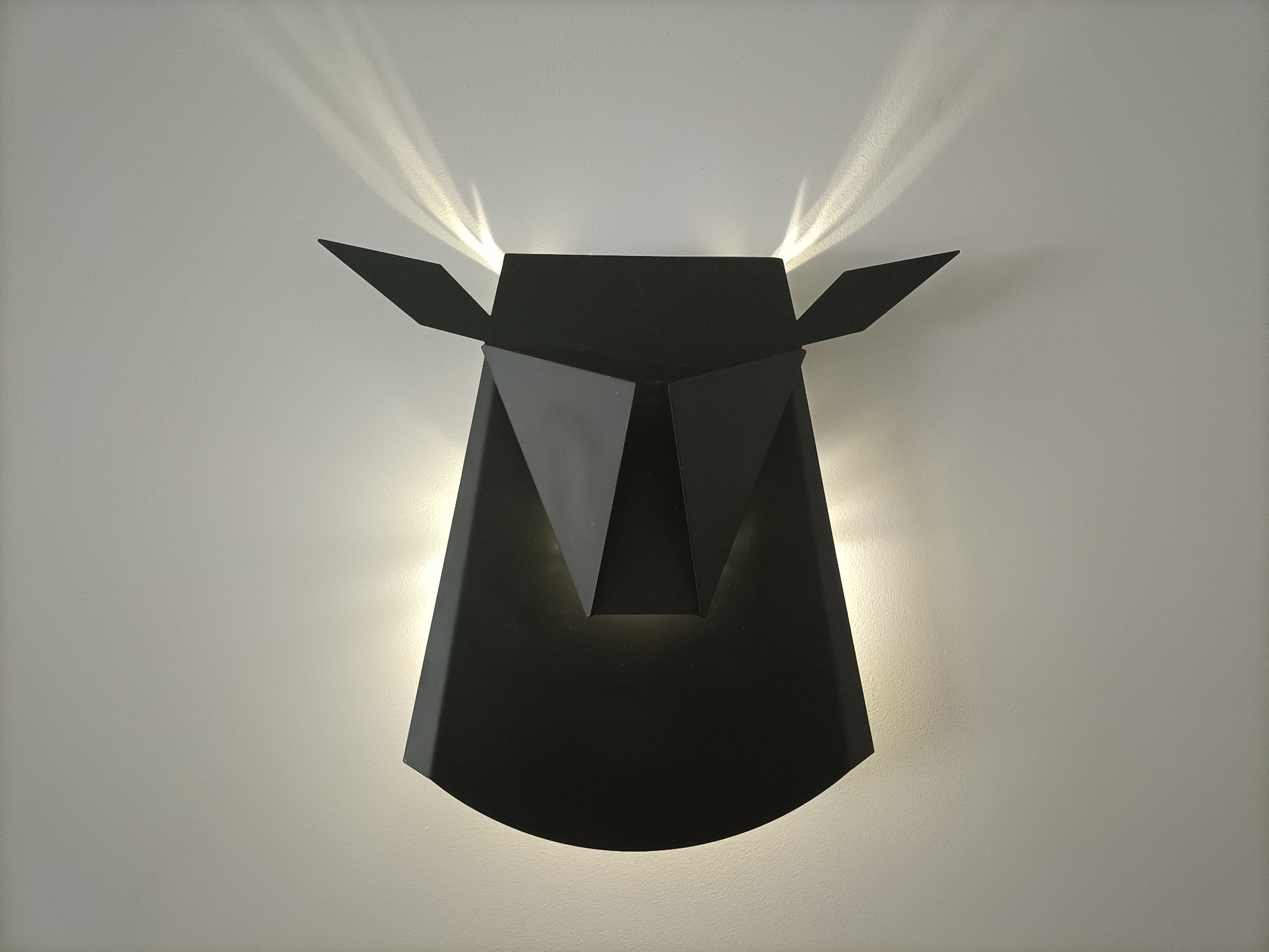 Czarny, geometryczny kinkiet ścienny w kształcie głowy zwierzęcia z oświetleniem skierowanym w górę i w dół.