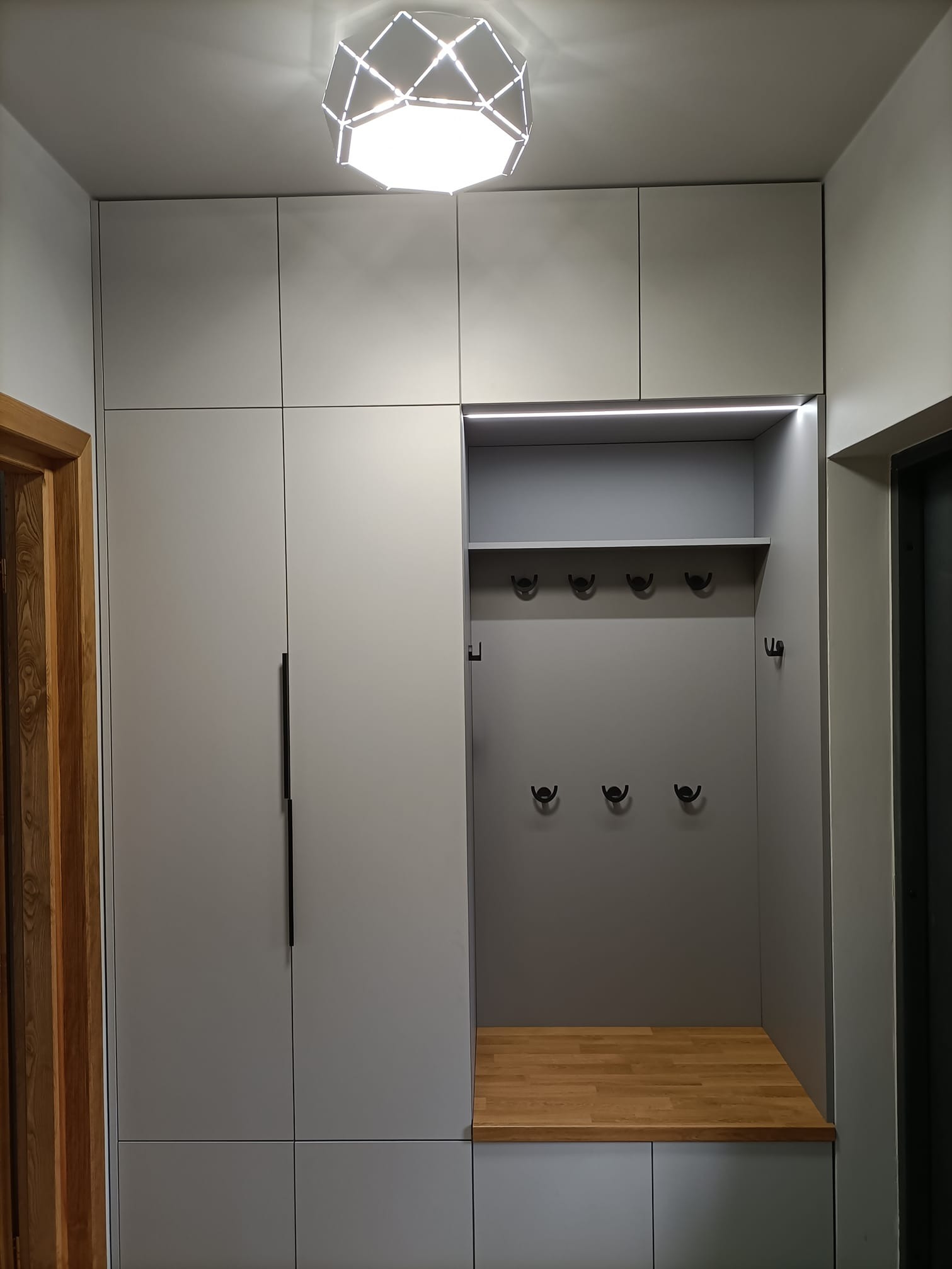 Szara zabudowa przedpokoju z szafkami, wieszakami i drewnianym siedziskiem, oświetlona lampą sufitową i listwą LED.