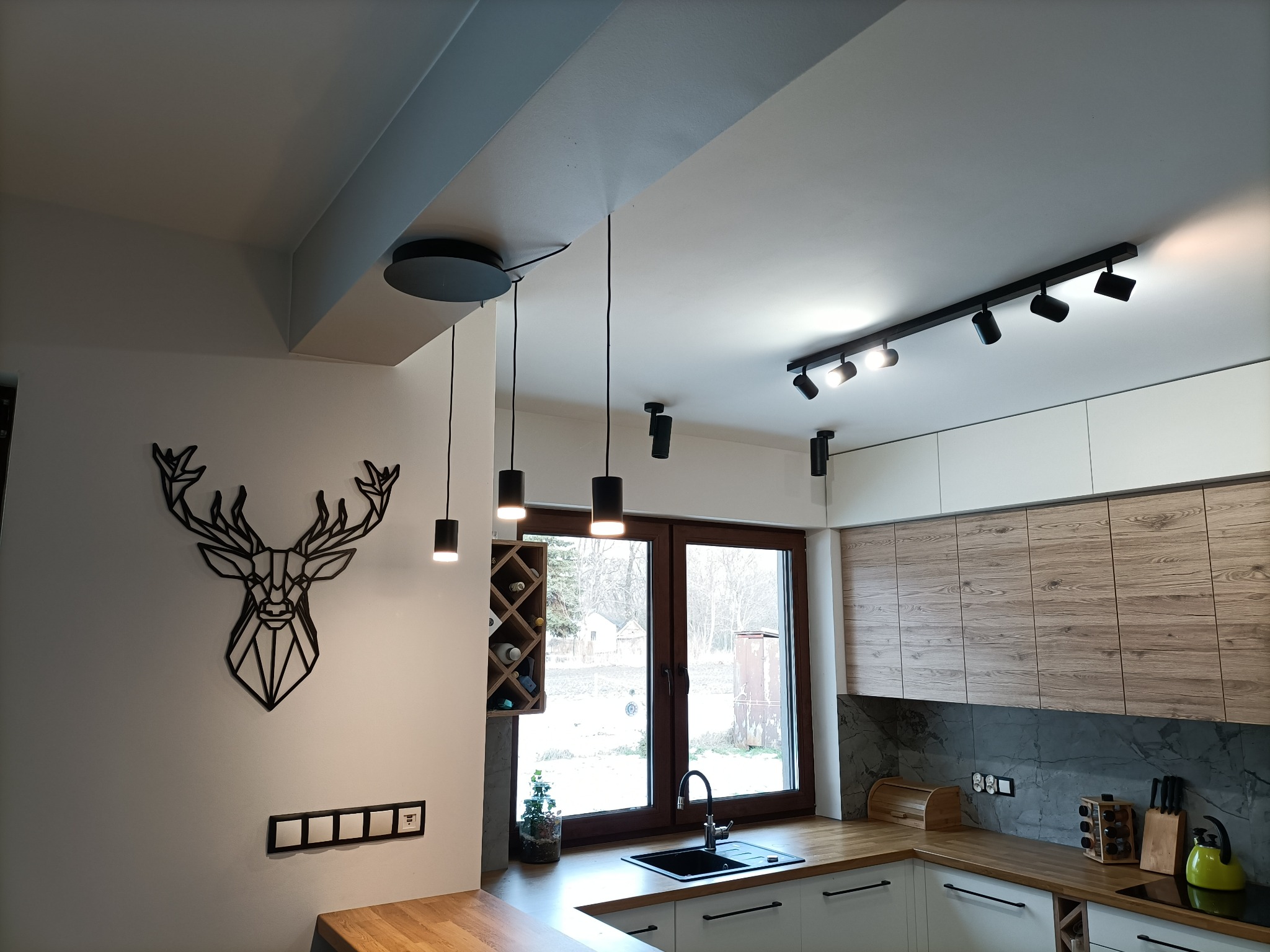 Nowoczesna kuchnia z drewnianym blatem, czarnym zlewem i baterią, oświetlona wiszącymi lampami oraz szynoprzewodem z reflektorami. Na ścianie dekoracja w kształcie głowy jelenia i włączniki światła.