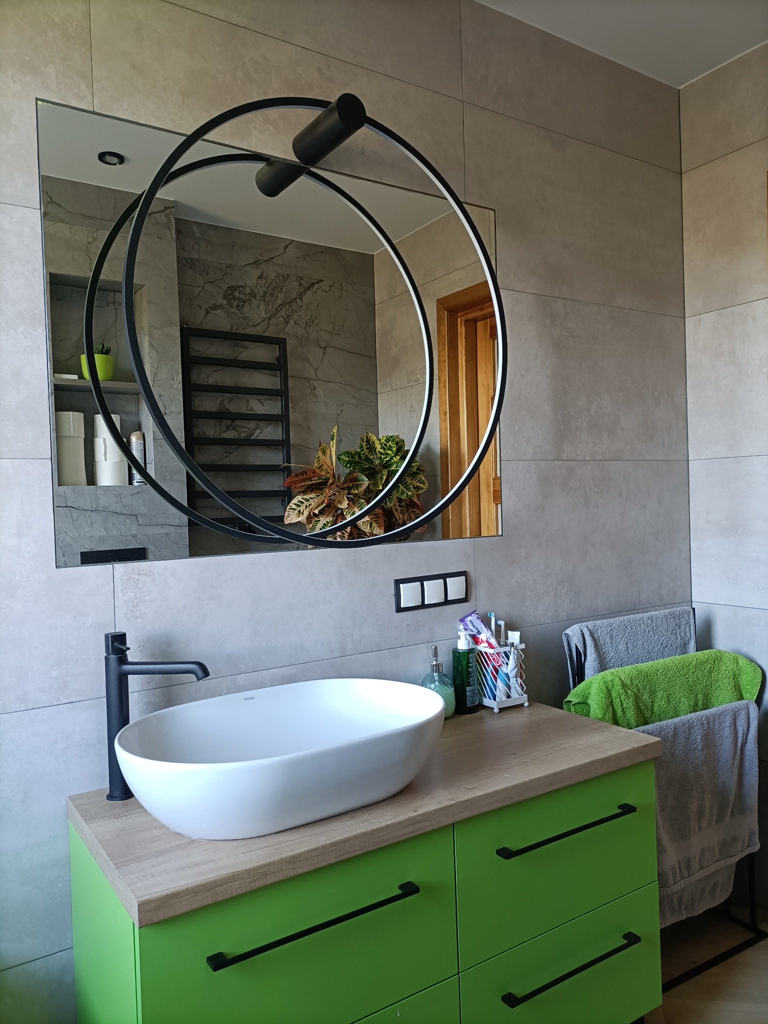Nowoczesna łazienka z umywalką nablatową, szafką w kolorze zielonego jabłka i okrągłym lustrem z podświetleniem LED, na tle szarych płytek imitujących kamień.
