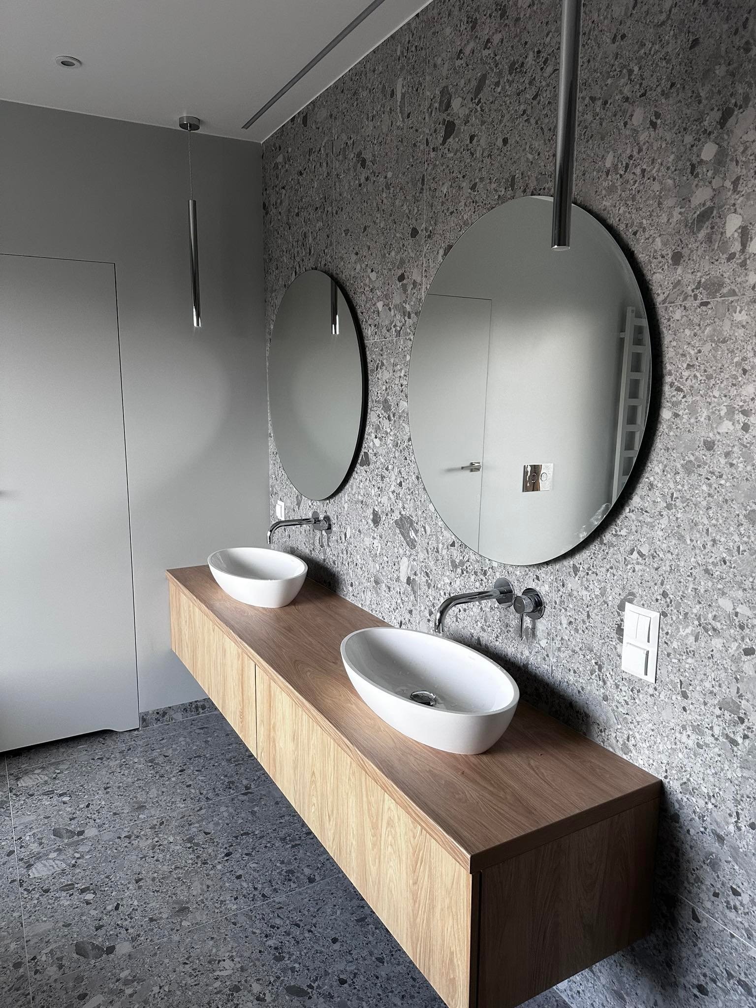 Nowoczesna łazienka z dwoma owalnymi umywalkami nablatowymi na drewnianej szafce, okrągłe lustra i wiszące lampy cylindryczne na tle ściany i podłogi z szarego lastryko.