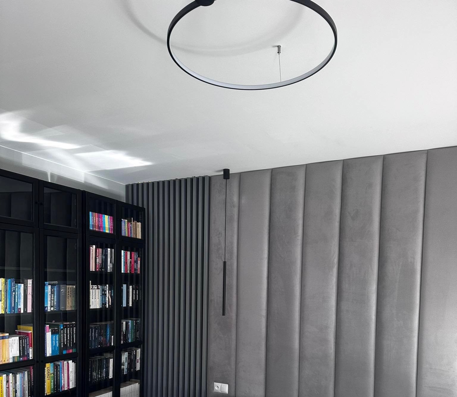 Nowoczesny salon z okrągłą lampą sufitową, minimalistyczną lampą wiszącą, panelem ściennym w odcieniach szarości oraz czarnym regałem z książkami.