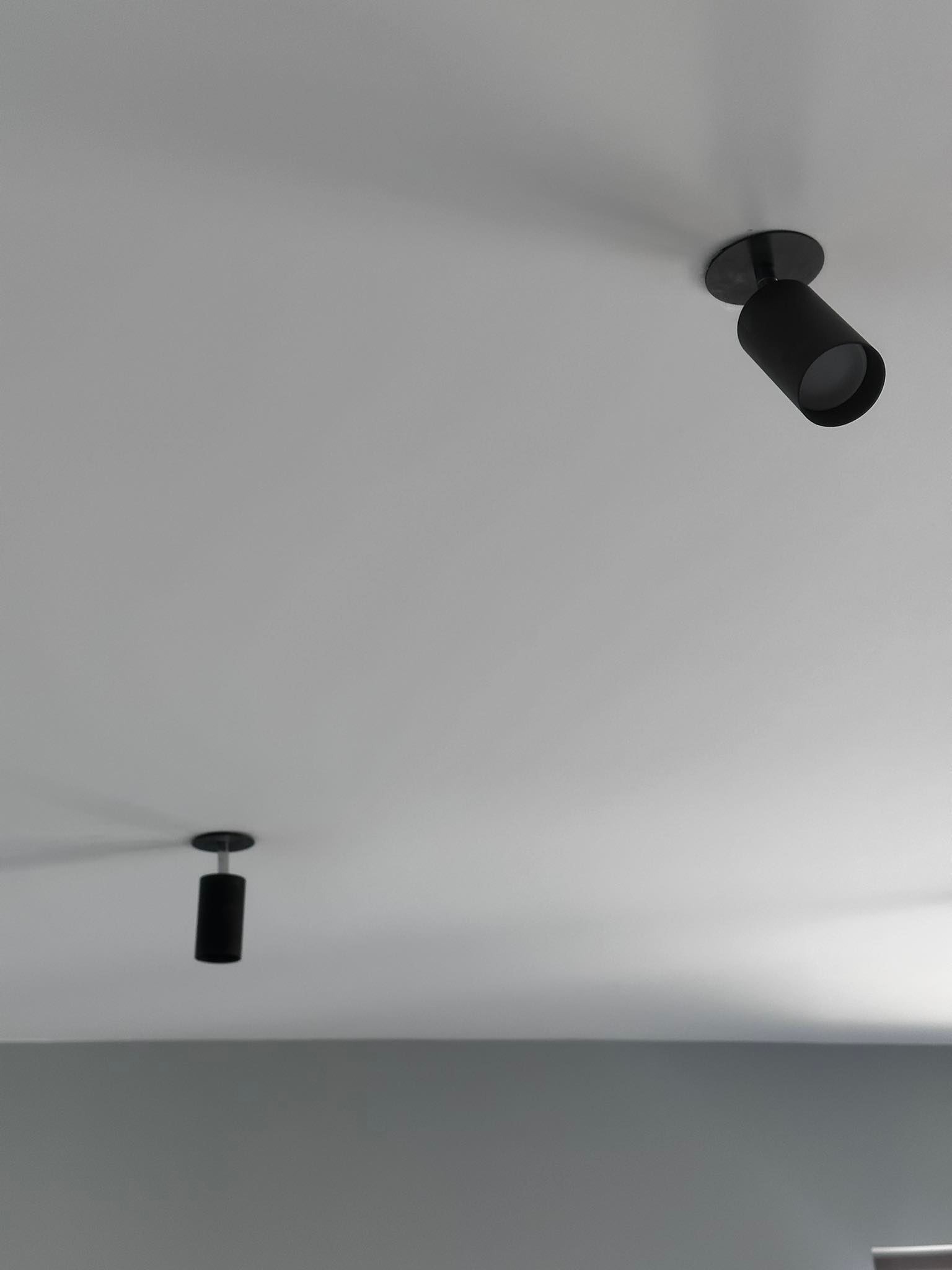 Dwa czarne reflektory punktowe zamontowane na białym suficie, oświetlające szarą ścianę w tle.
