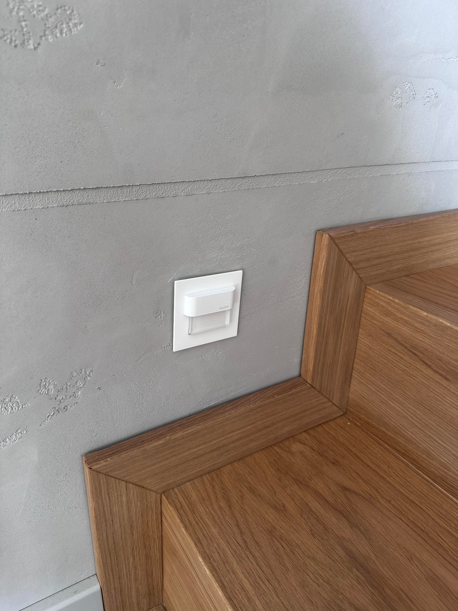 Biały czujnik ruchu na szarej ścianie strukturalnej, zamontowany przy drewnianej podłodze i listwie przypodłogowej.