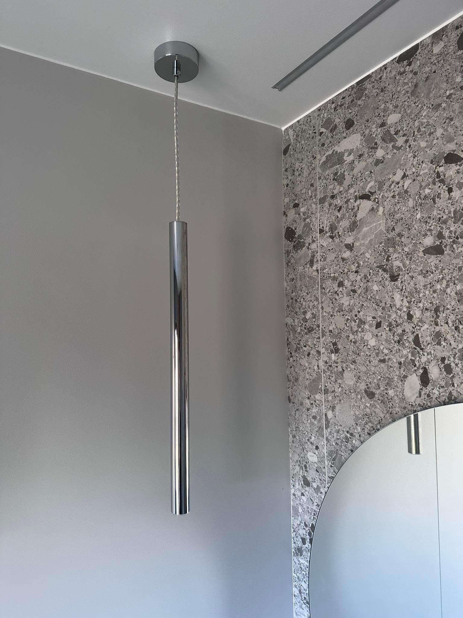 Minimalistyczna, chromowana lampa wisząca w łazience z szarą ścianą i mozaikową płytką, odbijająca się w okrągłym lustrze.