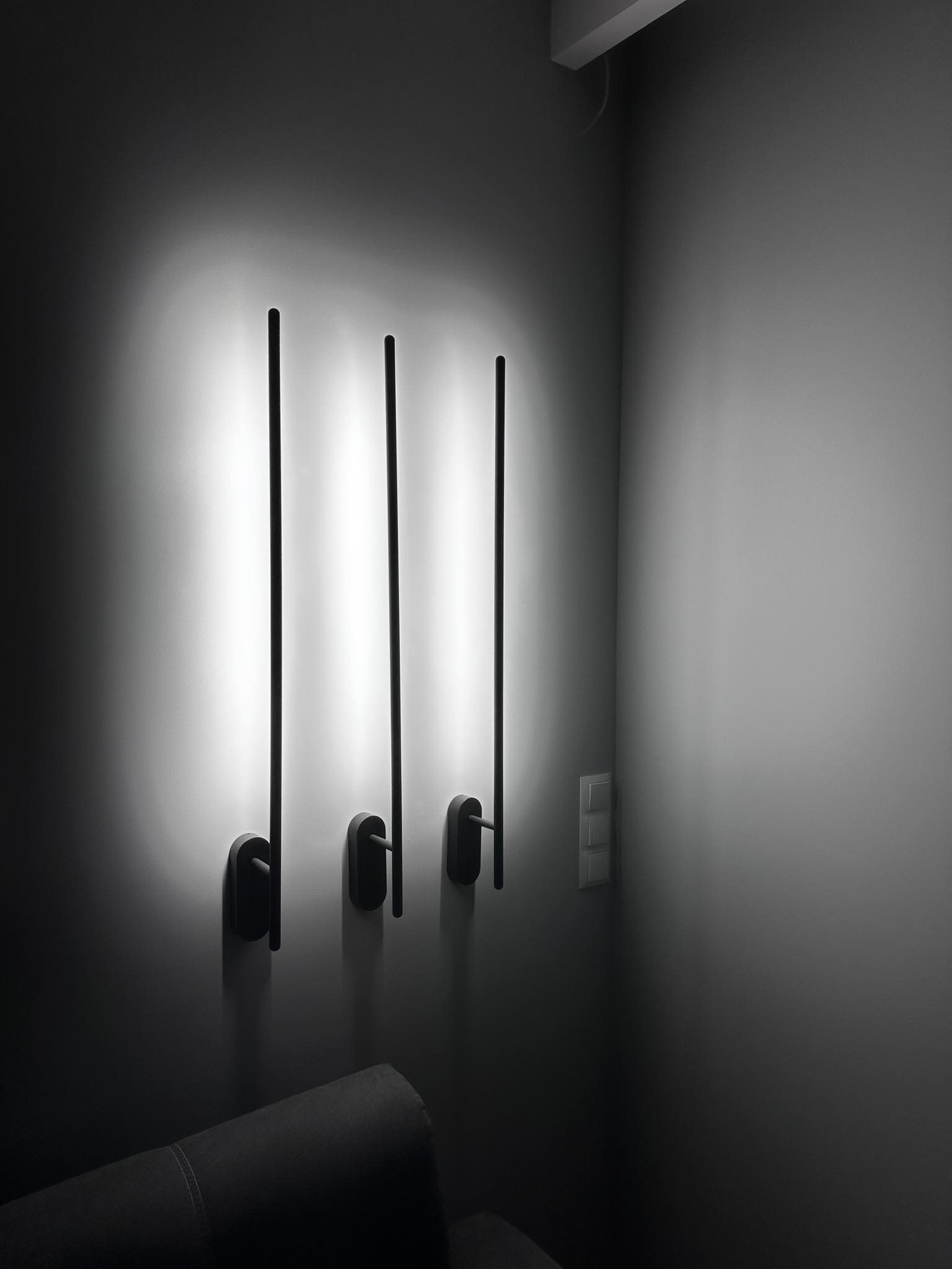 Minimalistyczne czarne lampy ścienne z pionowymi prętami, emitujące jasne, rozproszone światło na szarej ścianie w rogu pokoju, widoczny fragment szarej sofy.