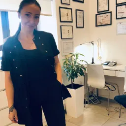 Kobieta w czarnym stroju medycznym, stojąca w gabinecie z certyfikatami na ścianie, biurkiem i rośliną doniczkową w tle.