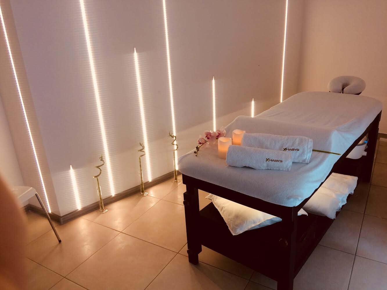 Przytulne wnętrze gabinetu z łóżkiem do masażu, białymi ręcznikami z logo, zapalonymi świecami, orchideą i dekoracyjnymi bambusami podświetlonymi pionowymi lampami LED.