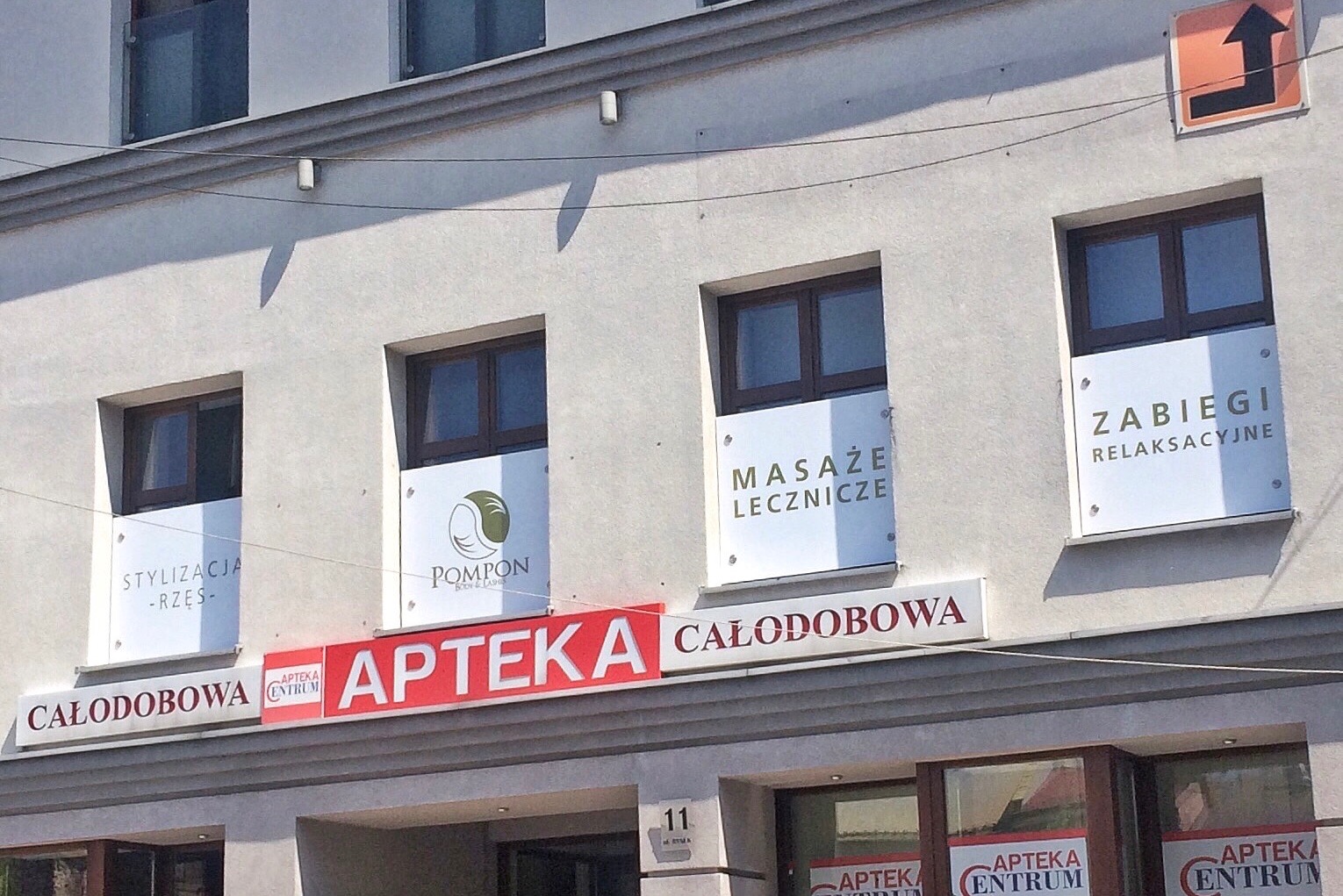 Elewacja budynku z szyldami reklamującymi usługi: stylizacja rzęs, masaże lecznicze, zabiegi relaksacyjne oraz logo salonu kosmetycznego Pompon Body & Lashes, nad apteką całodobową.