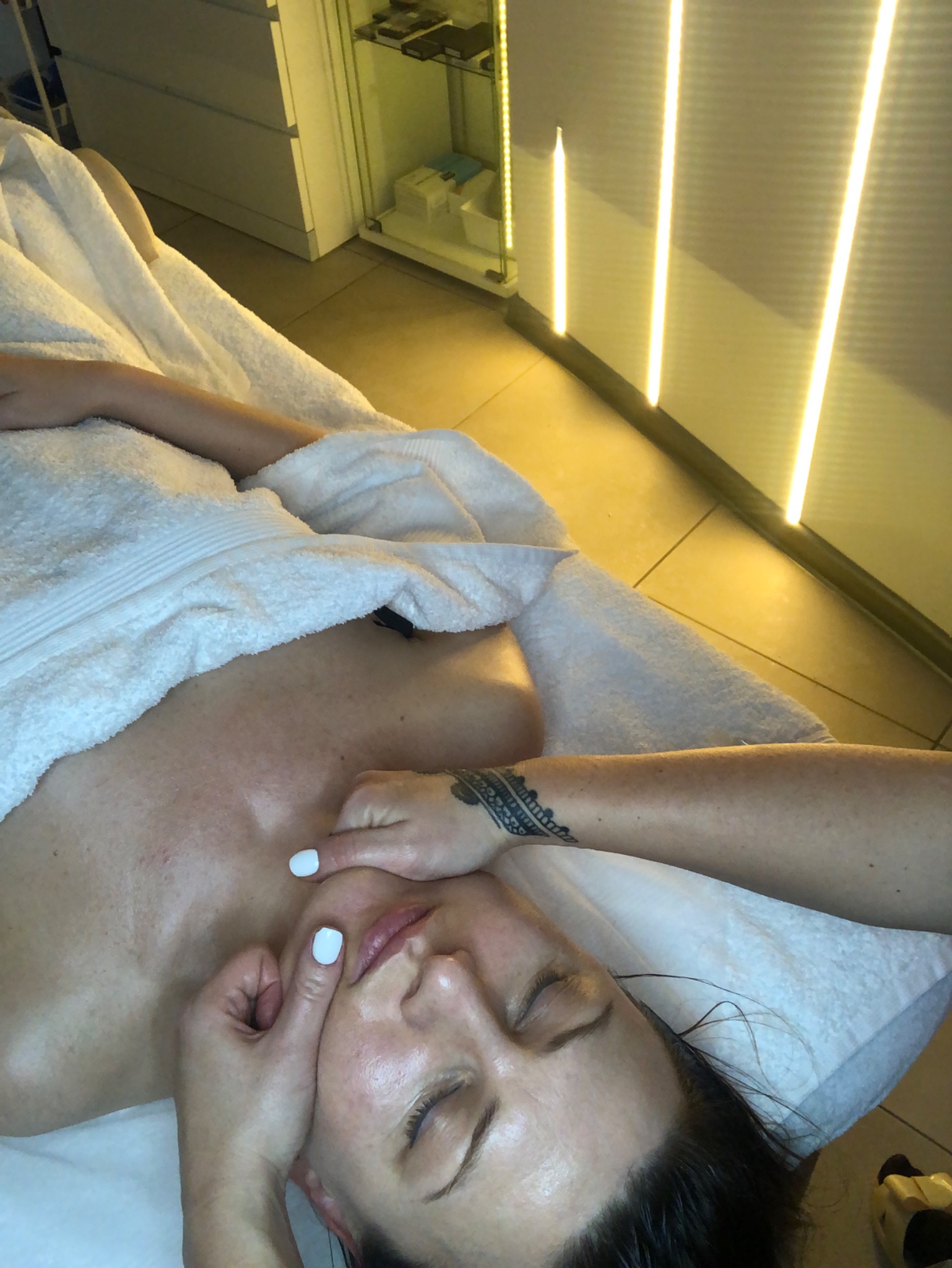 Kobieta leżąca na leżance pod białym ręcznikiem, podczas masażu twarzy wykonywanego przez osobę z tatuażem na przedramieniu, w gabinecie z oświetleniem LED.