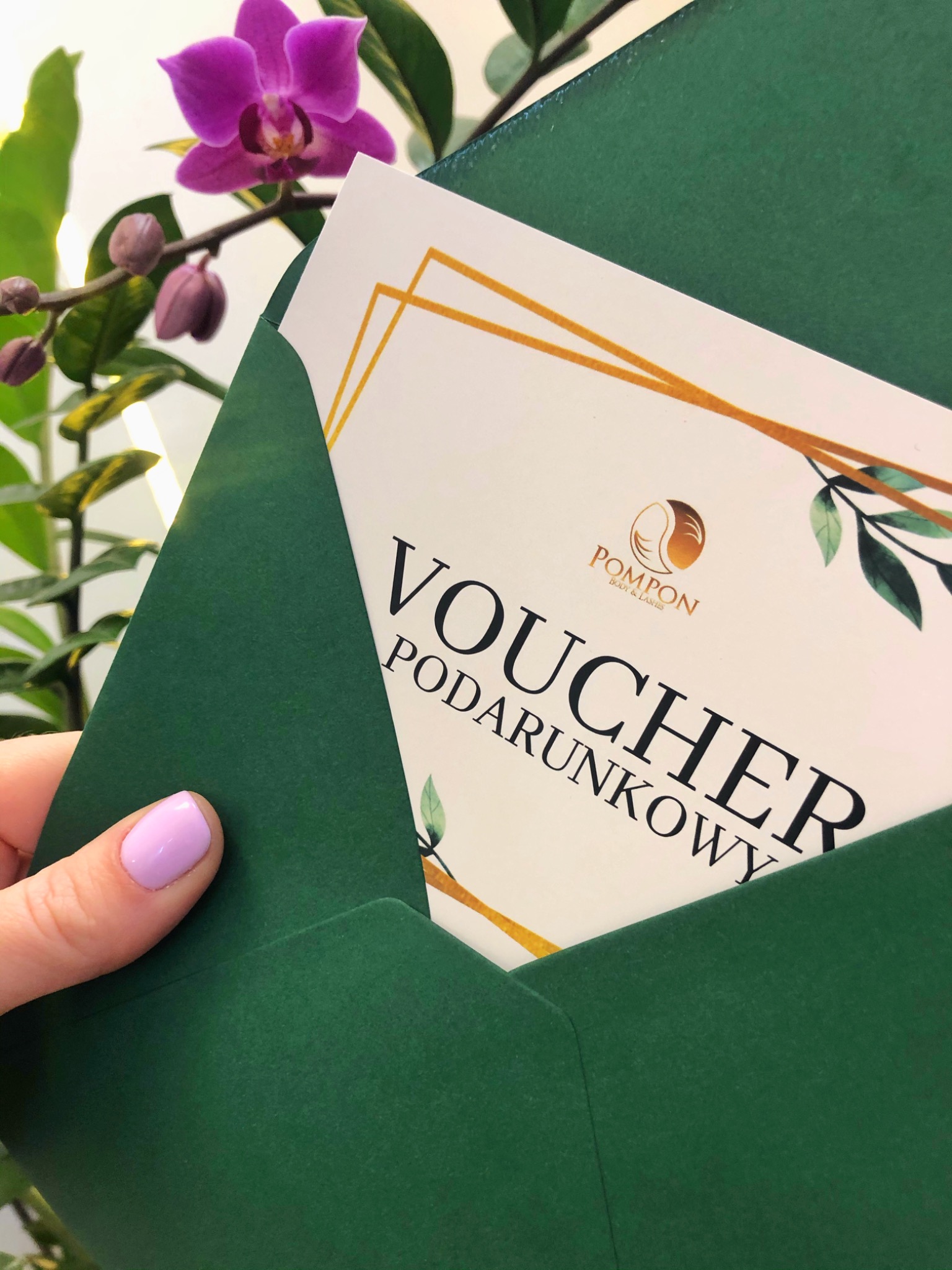Dłoń z delikatnym fioletowym manicure trzyma zieloną kopertę z voucherem prezentowym salonu kosmetycznego na tle rozmytej orchidei.