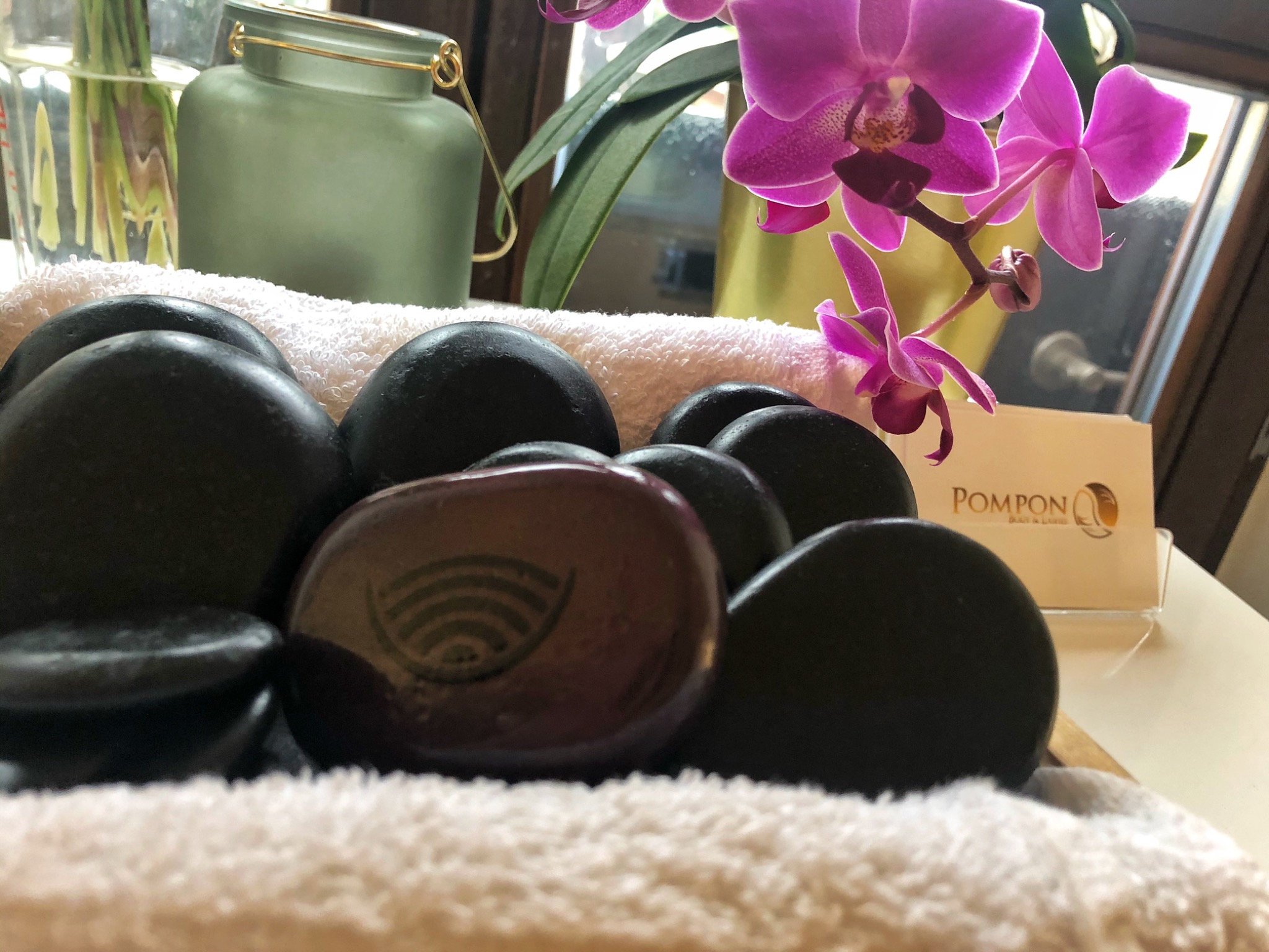 Zestaw czarnych, gładkich kamieni do masażu na białym ręczniku, z jednym brązowym kamieniem ozdobionym symbolem, obok wizytówka salonu kosmetycznego Pompon i kwitnąca orchidea.