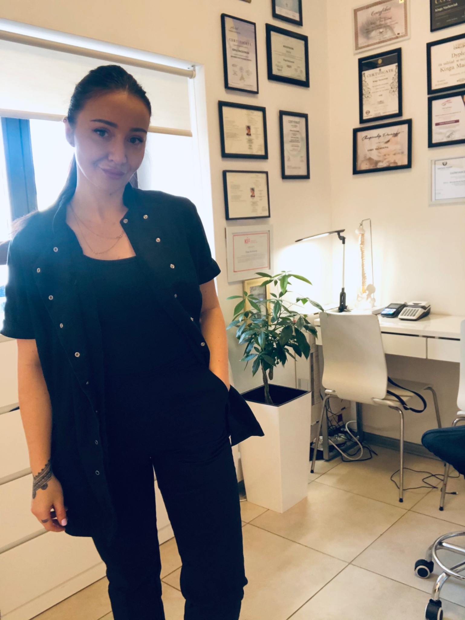 Kobieta w czarnym stroju medycznym, stojąca w gabinecie z certyfikatami na ścianie, biurkiem i rośliną doniczkową w tle.