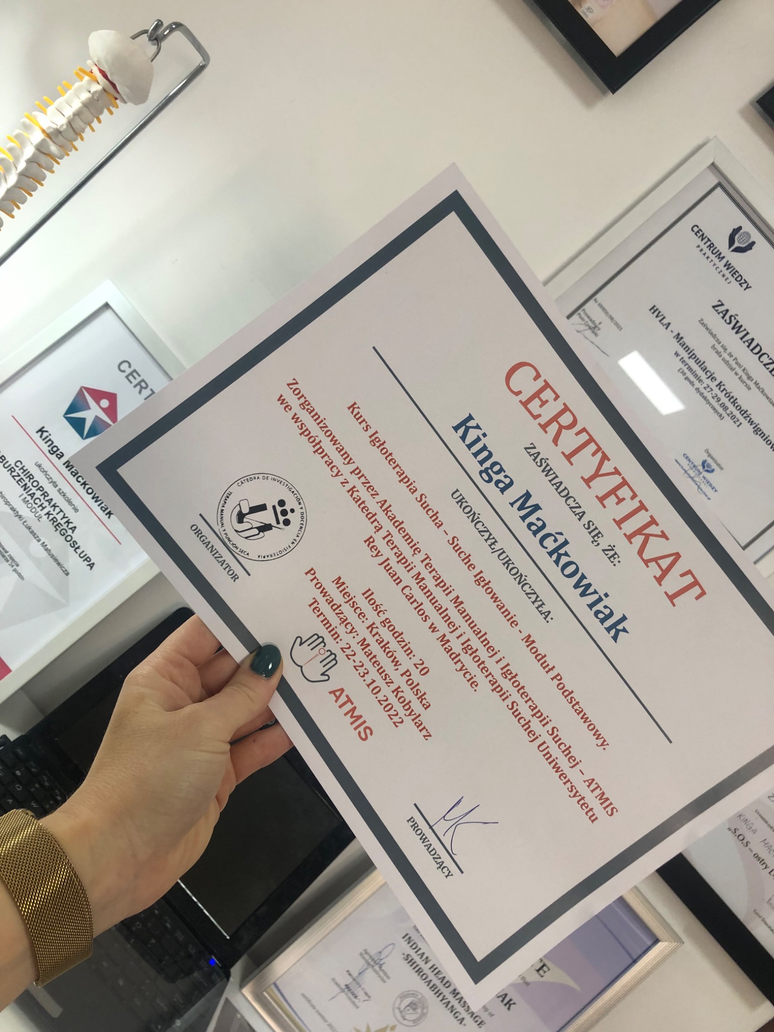 Kobieta trzyma certyfikat ukończenia kursu igłoterapii suchej Kingi Maćkowiak, widoczne inne certyfikaty na ścianie i model kręgosłupa w tle