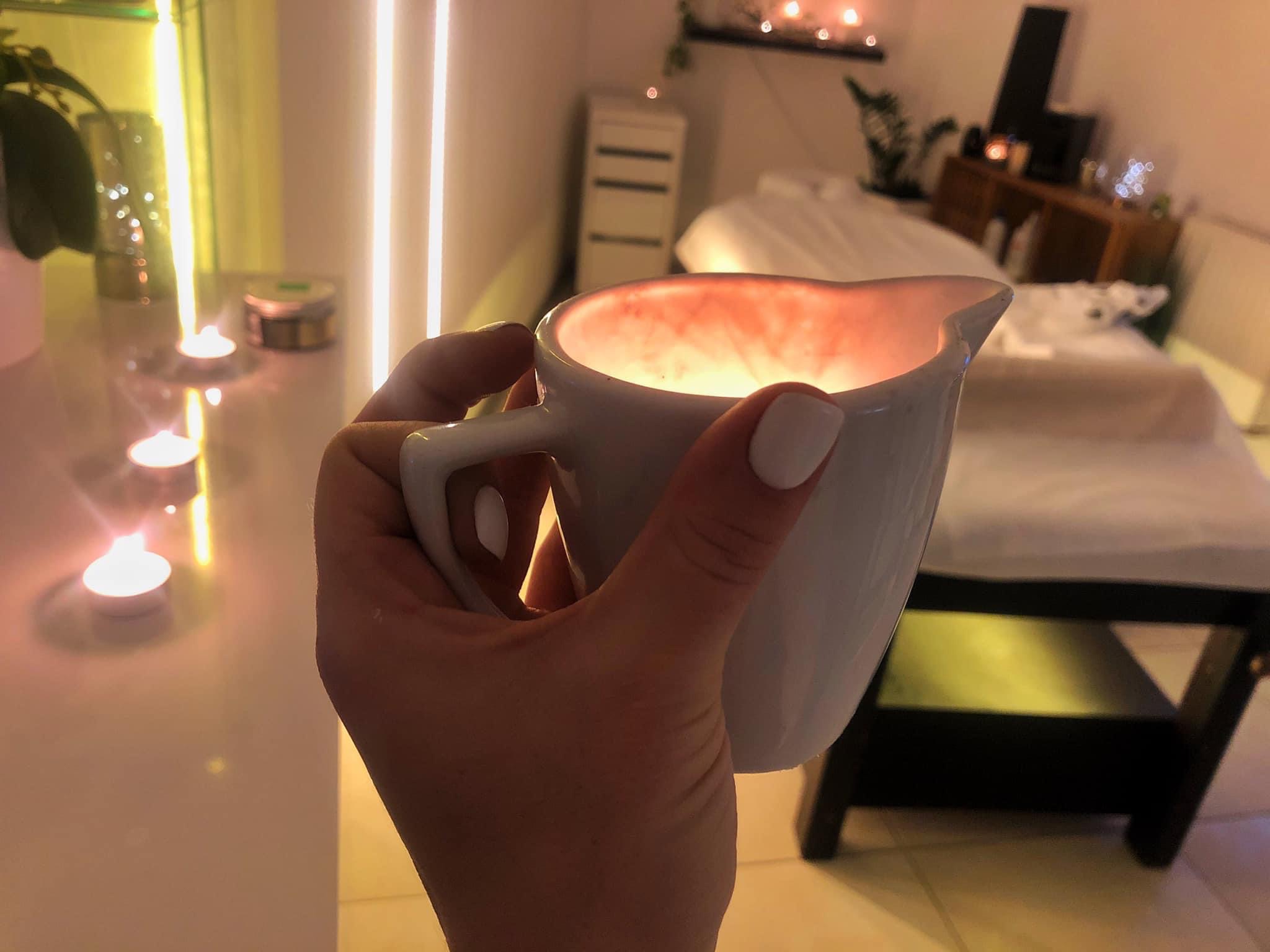 Dłoń trzymająca biały dzbanek z ciepłym olejkiem do masażu na tle gabinetu kosmetycznego z łóżkiem do masażu i świecami.