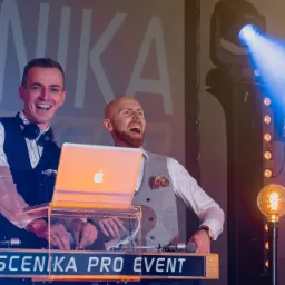 Dwóch uśmiechniętych mężczyzn w eleganckich kamizelkach obsługuje konsoletę DJ-ską z laptopem Apple, w tle profesjonalne oświetlenie sceniczne i dekoracyjne żarówki.