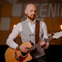 Muzyk z rudą brodą gra na akustycznej gitarze na scenie z oświetleniem punktowym i napisem 'Scenika Event' w tle.