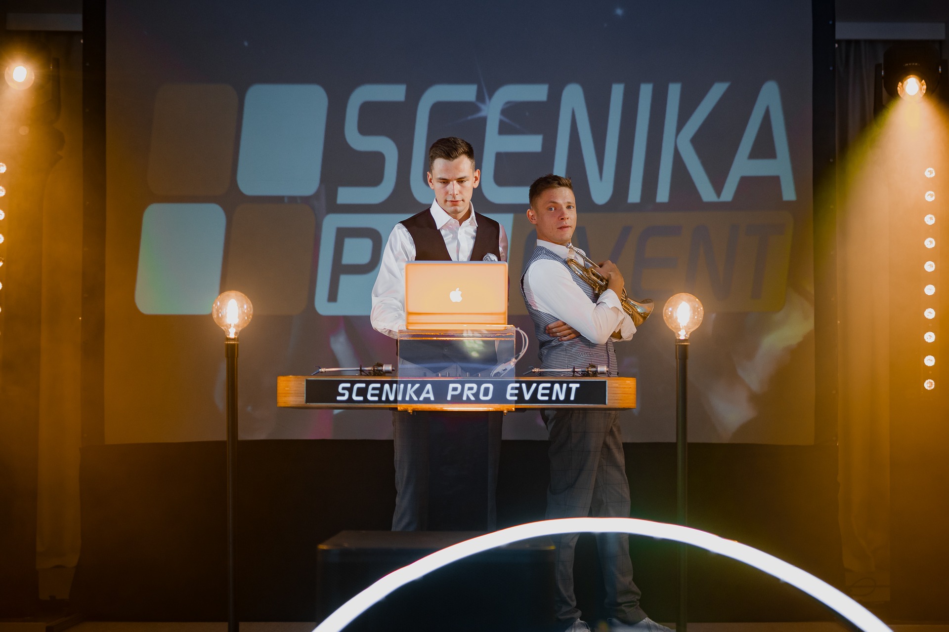 Dwóch mężczyzn w eleganckich strojach na scenie z laptopem i trąbką przed podświetlonym logo firmy eventowej, oświetlenie sceniczne i dekoracyjne żarówki.
