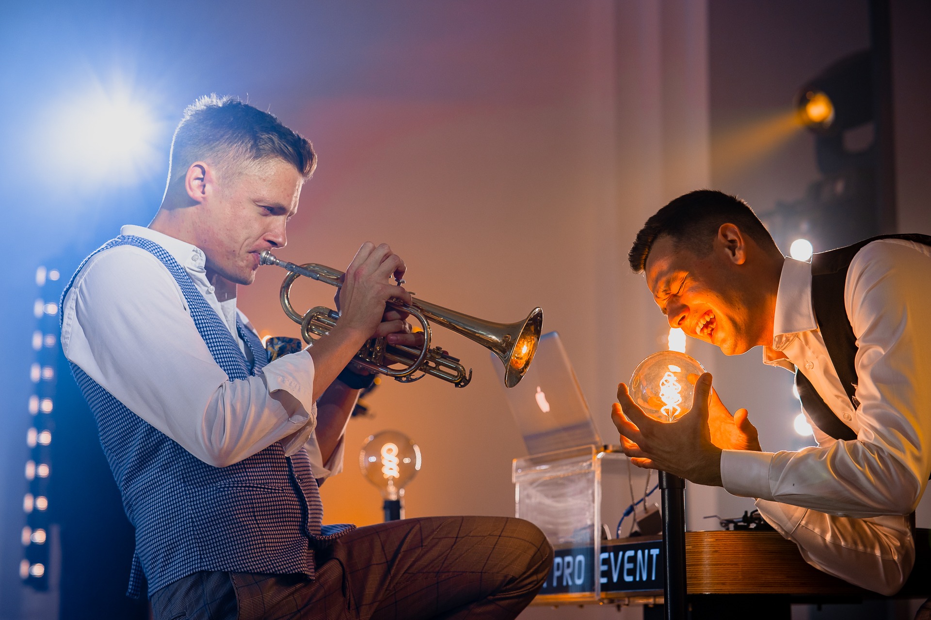 Muzyk grający na trąbce w kamizelce, obok mężczyzna trzymający świecącą żarówkę, oświetlenie sceniczne w tle.