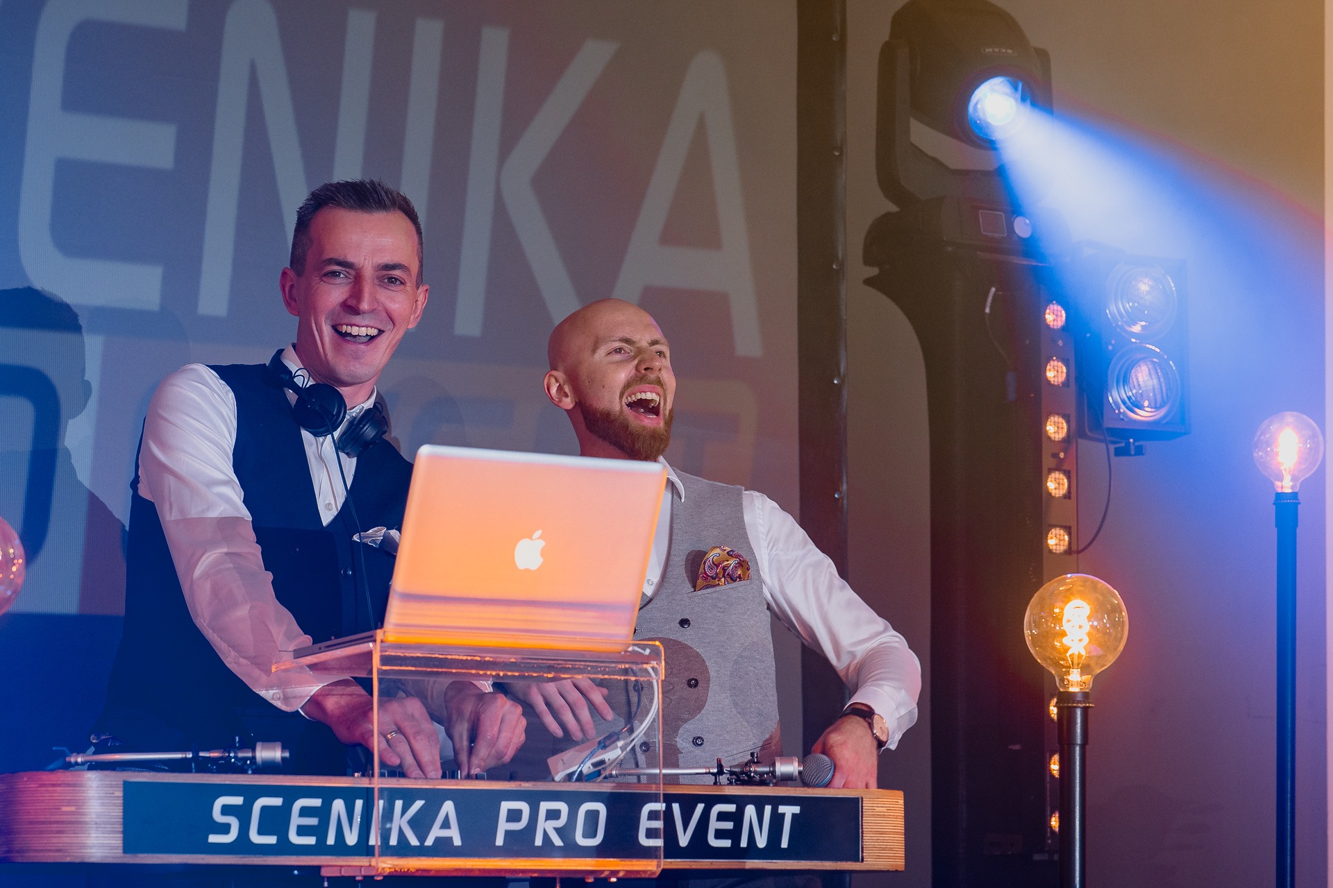 Dwóch uśmiechniętych mężczyzn w eleganckich kamizelkach obsługuje konsoletę DJ-ską z laptopem Apple, w tle profesjonalne oświetlenie sceniczne i dekoracyjne żarówki.