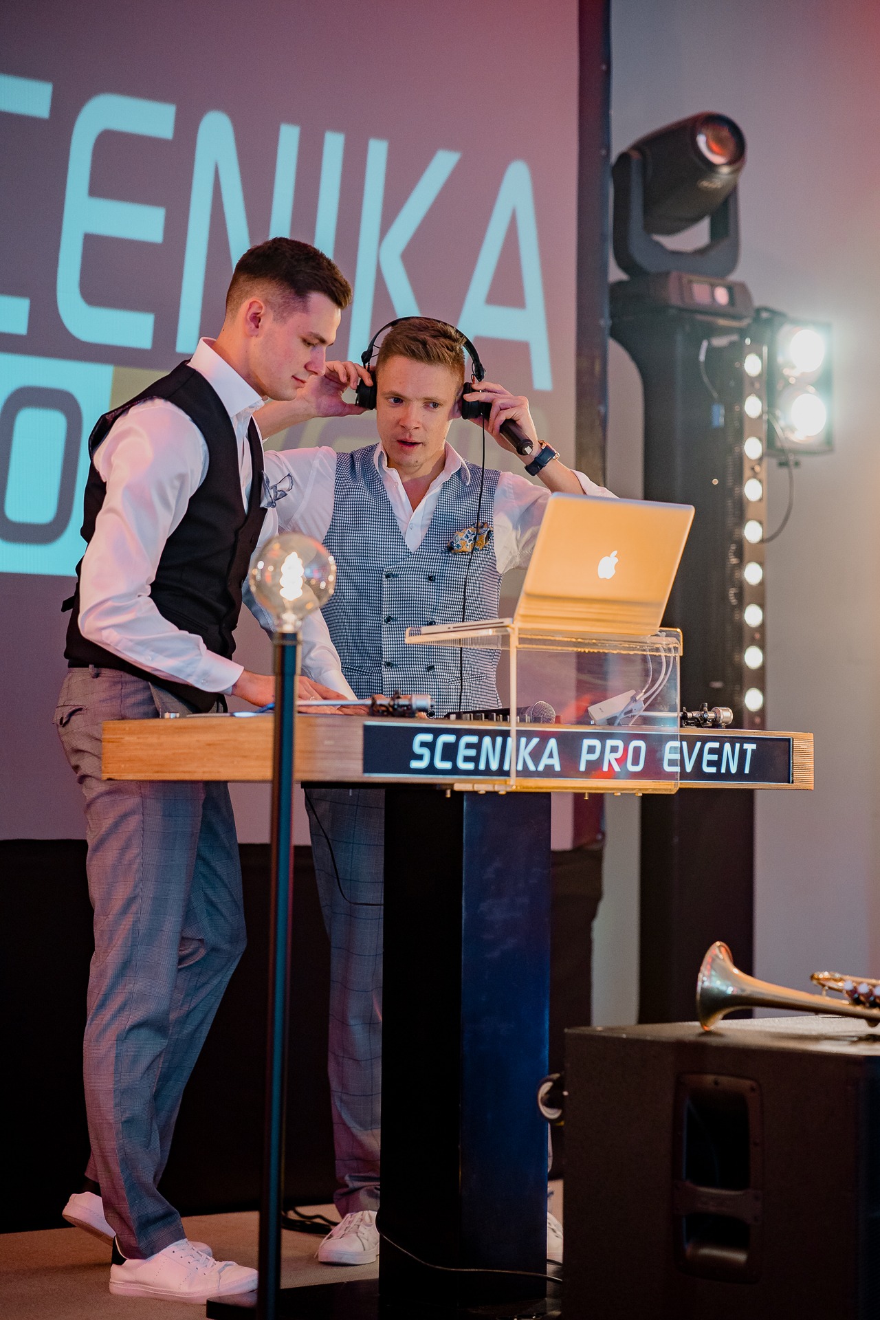 Dwóch mężczyzn w eleganckich strojach obsługuje konsoletę DJ-ską z laptopem i mikrofonem na tle oświetlonej sceny z napisem 'SCENIKA PRO EVENT', obok leży trąbka