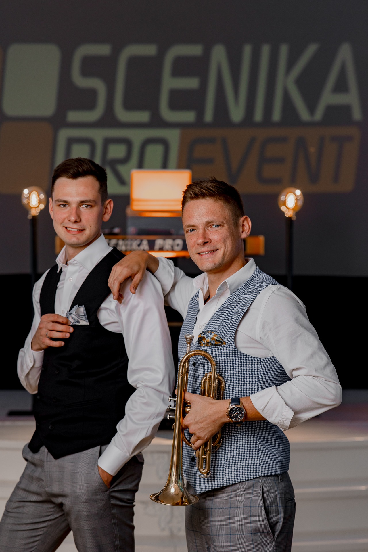 Dwóch elegancko ubranych mężczyzn pozuje z trąbką na tle sceny z logo 'SCENIKA PRO EVENT', oświetlonej żarówkami w stylu retro.