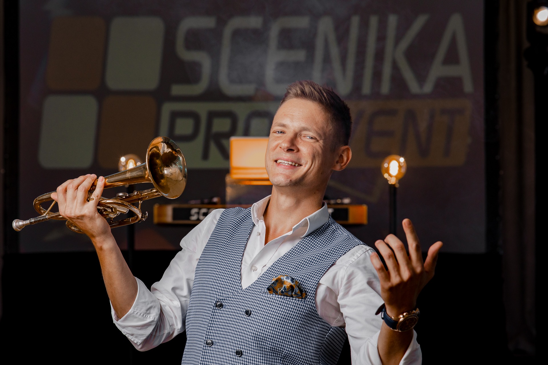 Uśmiechnięty trębacz w eleganckiej kamizelce prezentuje instrument na tle sceny z napisem 'SCENIKA PRO EVENT'.