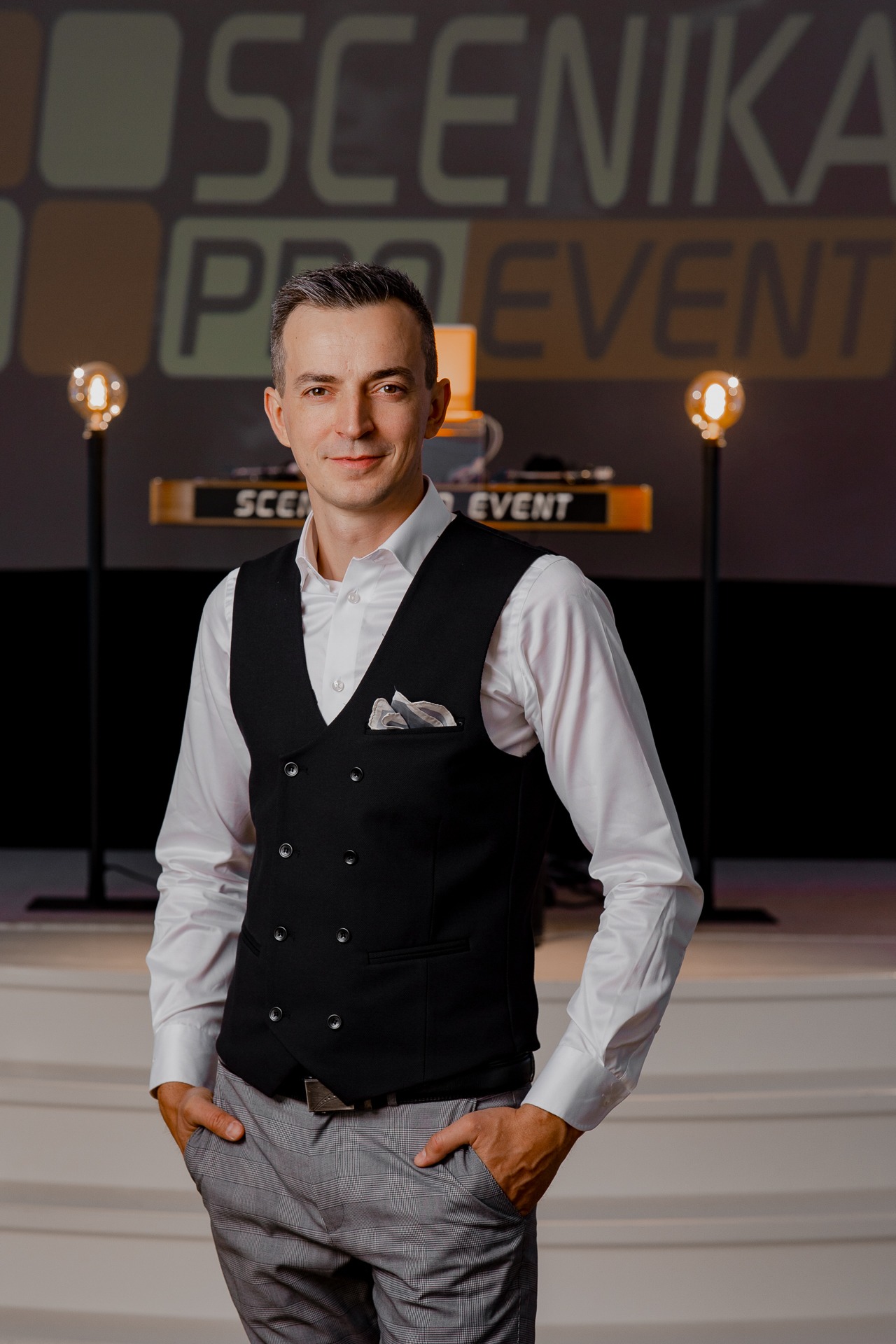 Mężczyzna w eleganckiej kamizelce na tle sceny z oświetleniem i napisem 'SCENIKA PRO EVENT'.