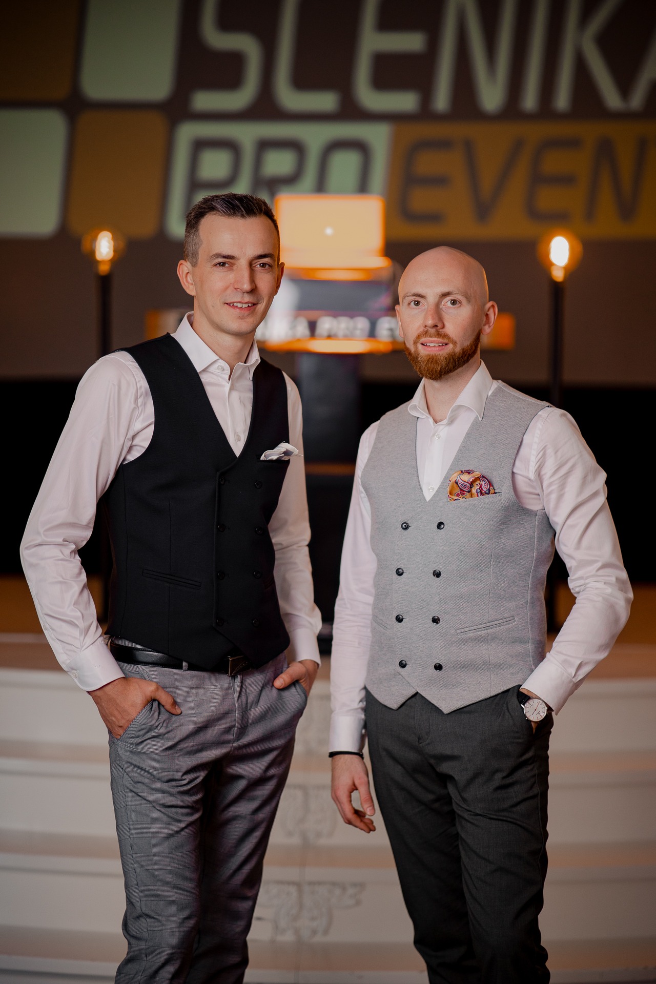 Dwóch elegancko ubranych mężczyzn w kamizelkach, pozujących na tle sceny z logo firmy eventowej, jeden z chusteczką w kieszonce kamizelki.