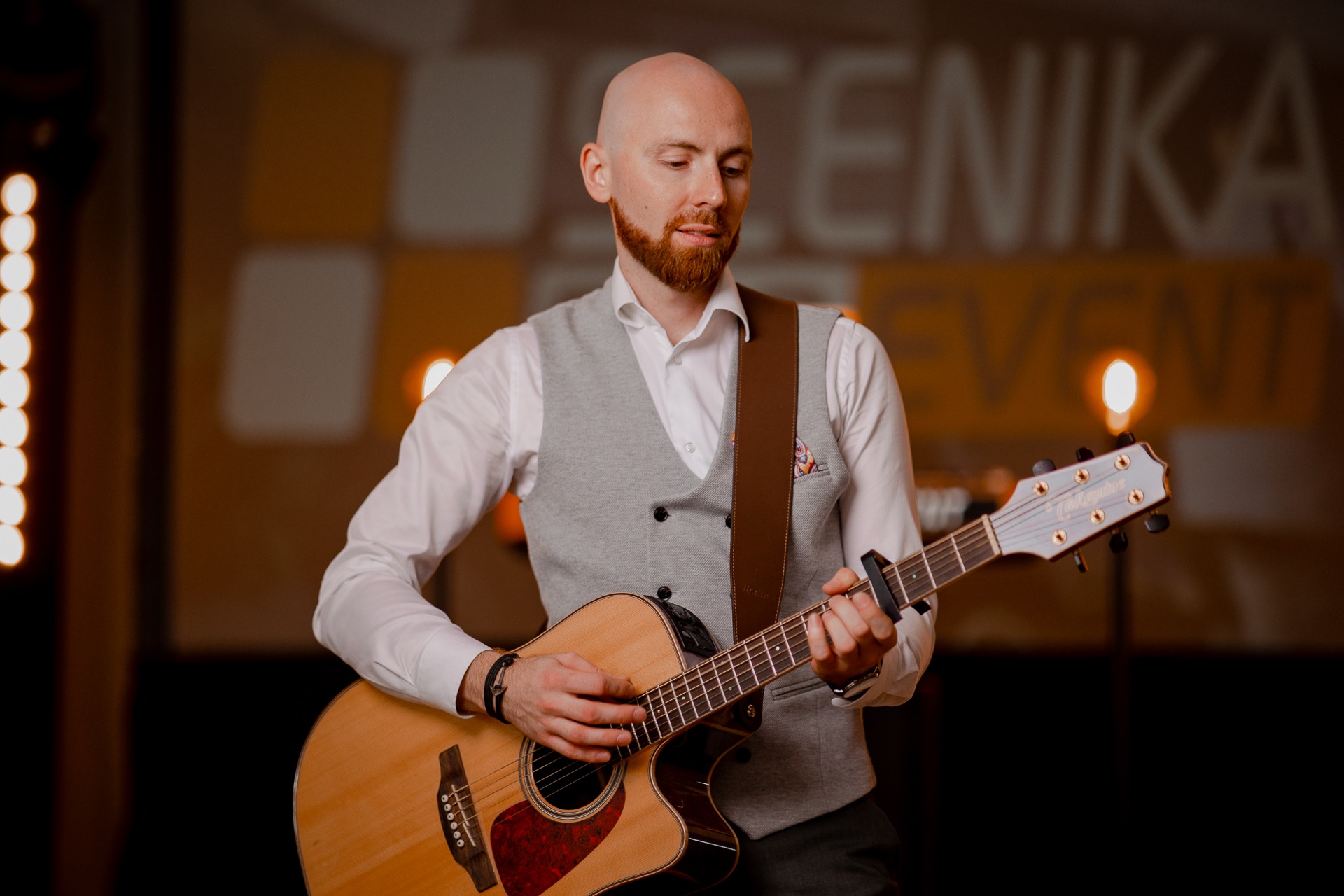 Muzyk z rudą brodą gra na akustycznej gitarze na scenie z oświetleniem punktowym i napisem 'Scenika Event' w tle.