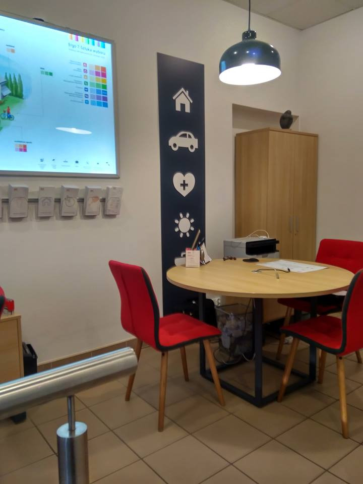 Wnętrze biura z okrągłym stołem i czerwonymi krzesłami, na ścianie grafika z ikonami domu, samochodu, serca i słońca, obok monitor z kolorową grafiką.