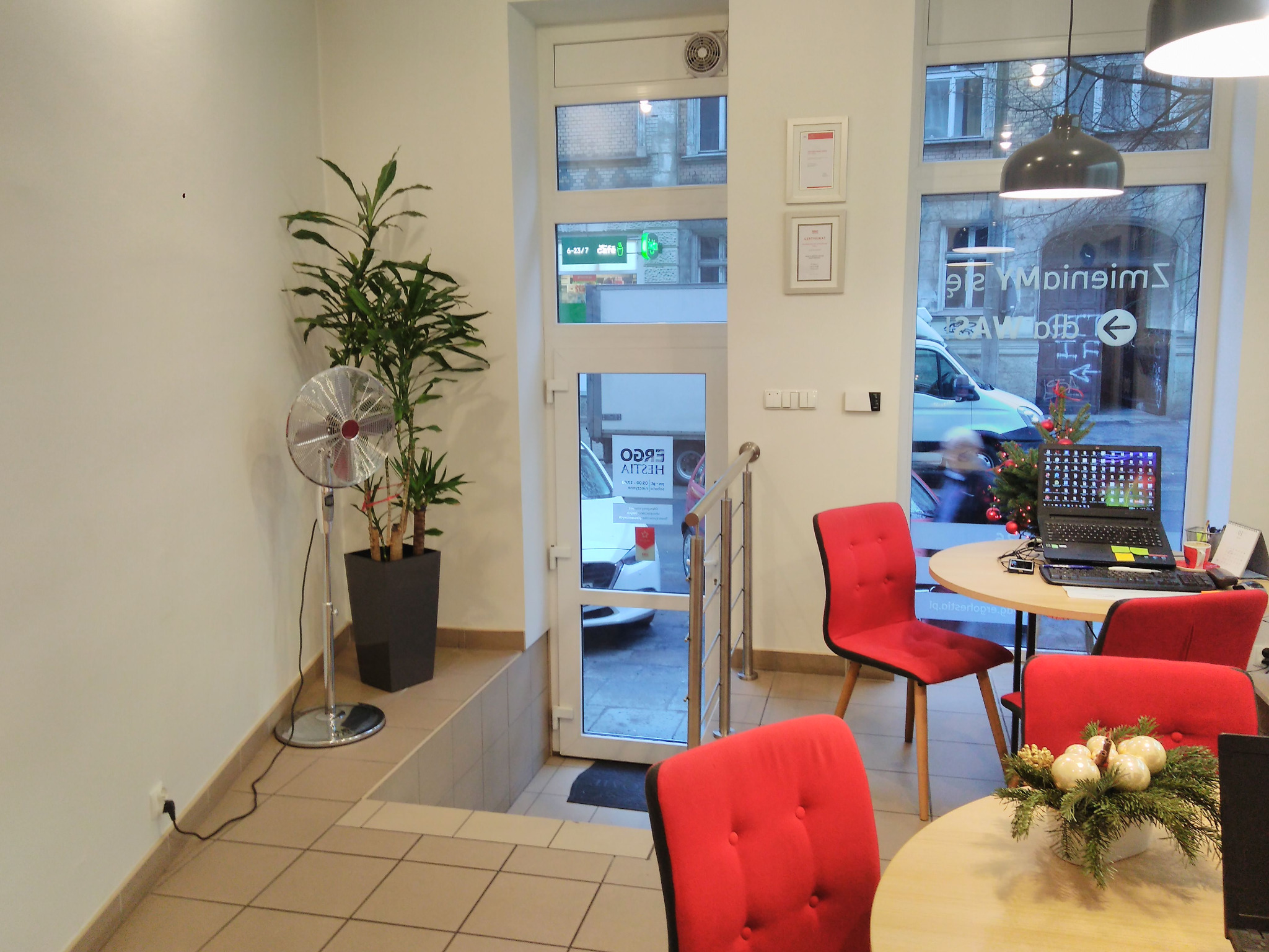 Jasne biuro z czerwonymi krzesłami, biurkami i komputerami, widok na ulicę przez okno z odbiciem napisu i świąteczną dekoracją.
