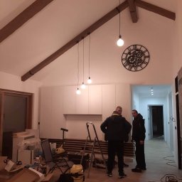 Natalii kuzyna - Wnętrze w trakcie prac wykończeniowych: drewniane belki stropowe, wiszące lampy, duży zegar na ścianie, widoczny bałagan i narzędzia, ekipa remontowa w tle.