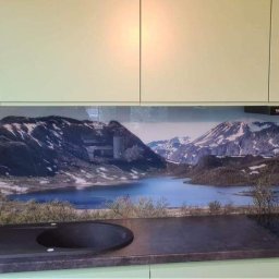 Natalii kuzyna - Szklana ściana nad blatem kuchennym z motywem górskiego krajobrazu z jeziorem, zamontowana między jasnymi szafkami a blatem z czarnym zlewem i płytą grzewczą.